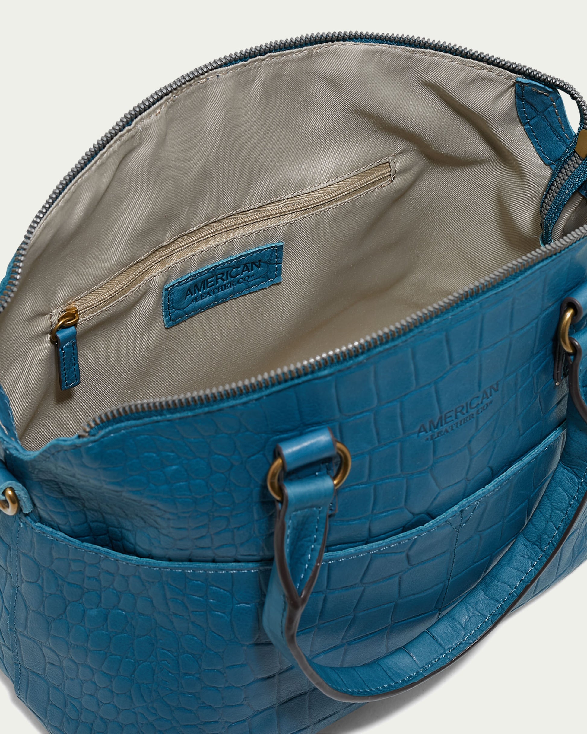 A teal-colored Carrie Dome Satchel from American Leather Co. with a crocodile skin pattern is open, revealing its interior. Inside, the bag features a beige lining, a zippered pocket, and a brand label that reads "Genuine American Leather." The handles are short and attached with metal rings; an adjustable crossbody strap is included.