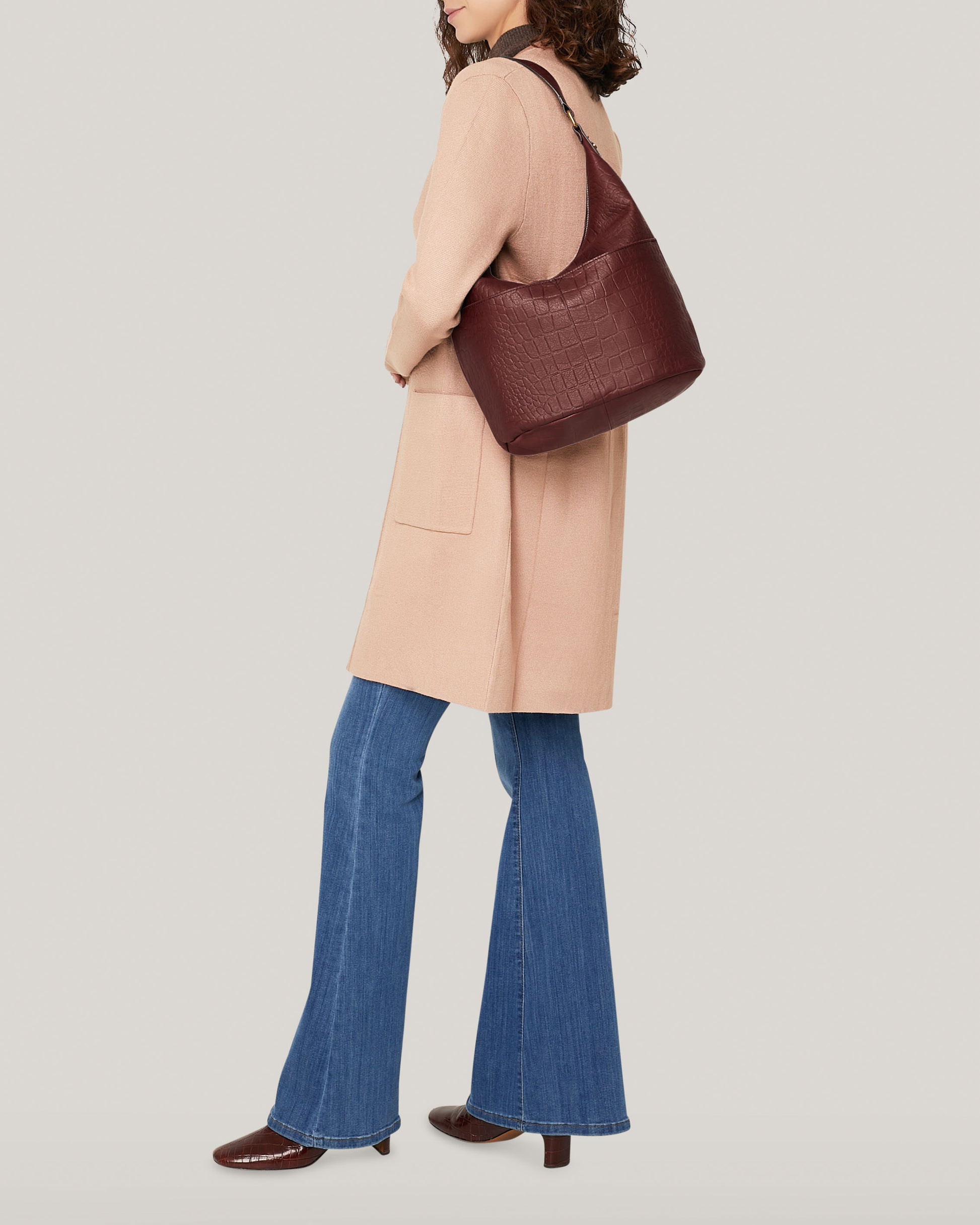 A person with curly hair is wearing a long beige coat, blue flare jeans, and brown heeled boots. They're carrying the Carrie Hobo by American Leather Co. over their shoulder, standing against a plain light background.