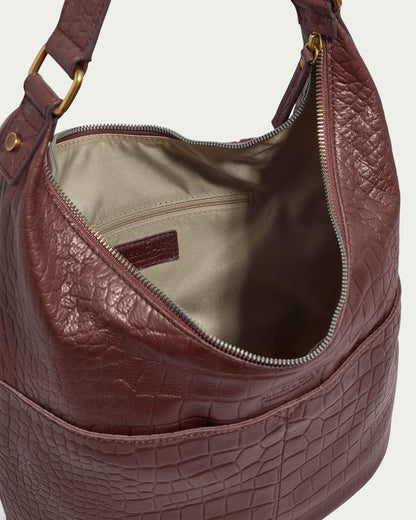 The Carrie Hobo by American Leather Co. is a brown bag made from American leather, showcasing a textured crocodile pattern. Its interior is lined with beige fabric, and it features a single shoulder strap with metal ring attachments along with a zip closure for added style and security.