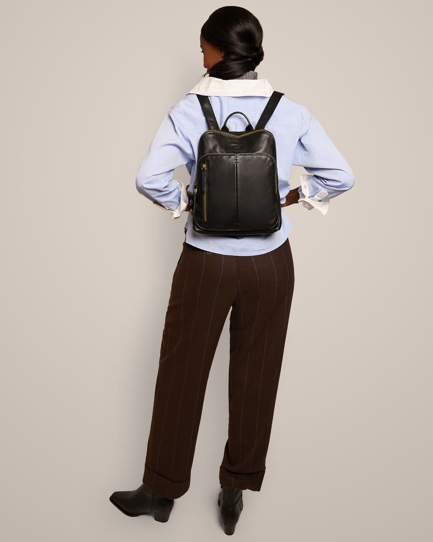 A person with long hair in a low bun is wearing a blue shirt and brown striped pants, facing away. They are sporting the Cleveland Backpack by American Leather Co., which features adjustable back straps, along with black ankle boots. The background is gray.