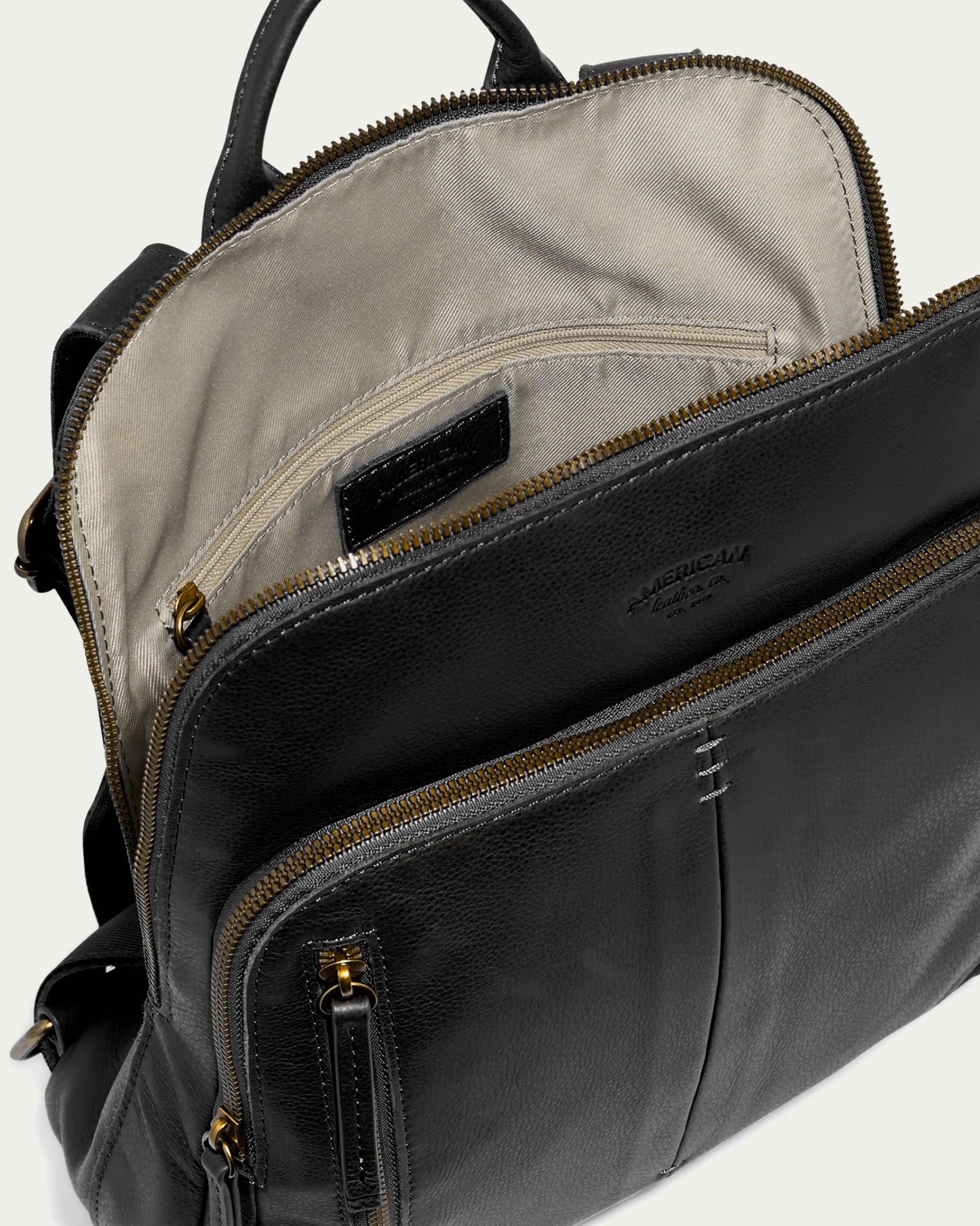 Discover the Cleveland Backpack from American Leather Co., an elegant black leather bag featuring adjustable back straps and multiple compartments with gold zippers. Made from genuine American leather, it boasts a beige interior with a visible pocket inside. It also includes two front pockets and a top handle for versatile carrying options.