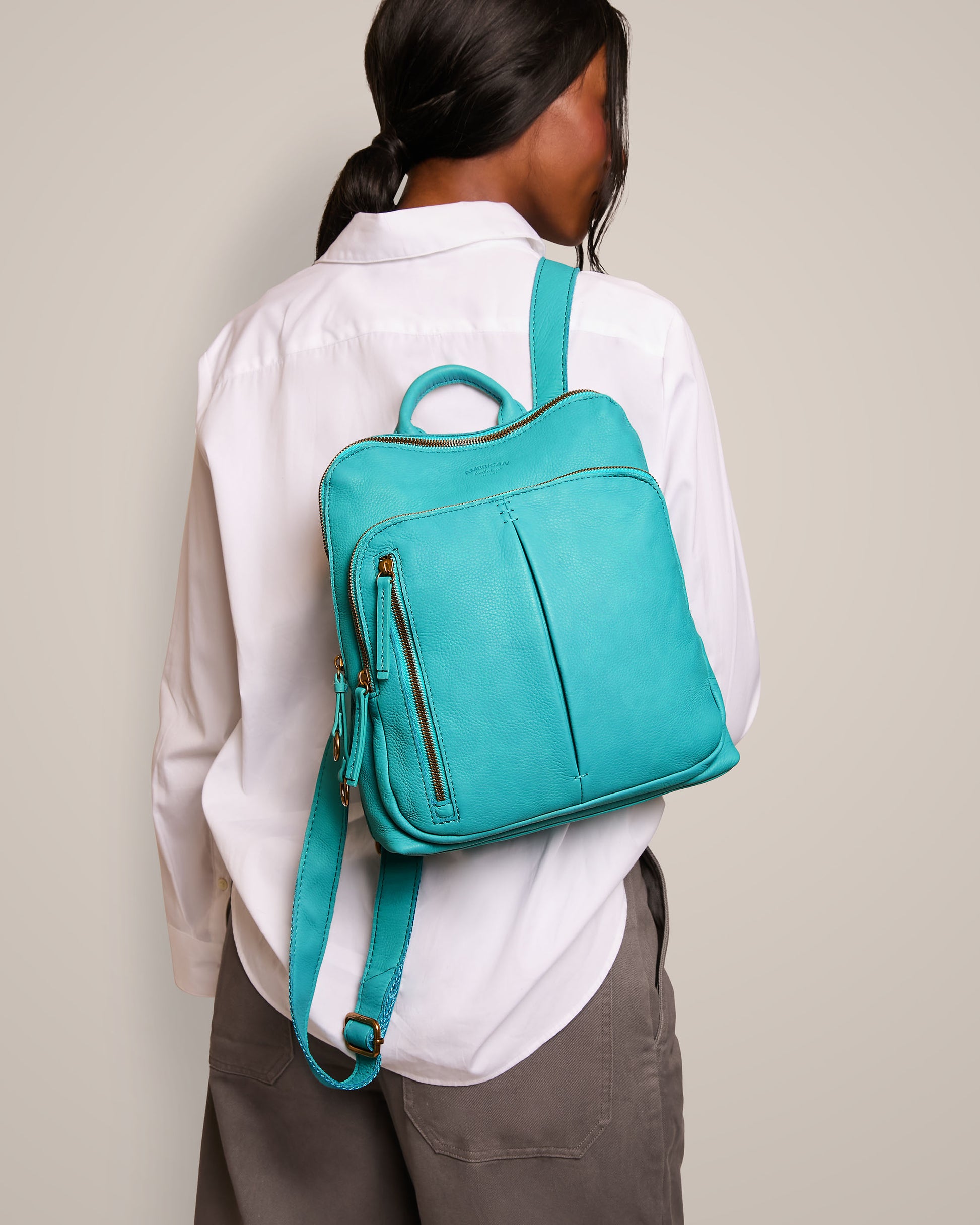 A person with long hair, dressed in a white shirt and gray pants, is seen from the back. They are carrying the Cleveland Backpack by American Leather Co., which is turquoise with adjustable back straps and features a small zipper pocket on the side.