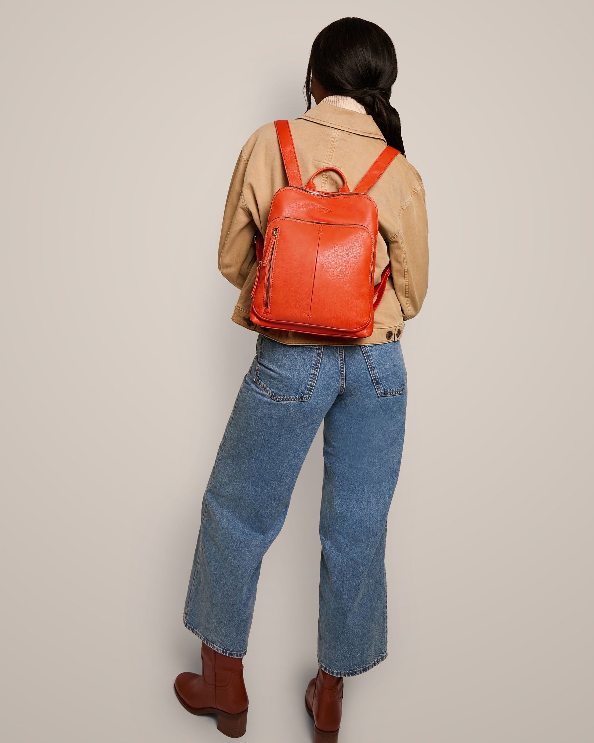 A person in a tan jacket and blue jeans stands facing away, showcasing the stylish Cleveland Backpack by American Leather Co., a bright red leather backpack purse. They have long hair and are wearing brown ankle boots. The plain background enhances the focus on the outfit and genuine leather Cleveland Backpack.