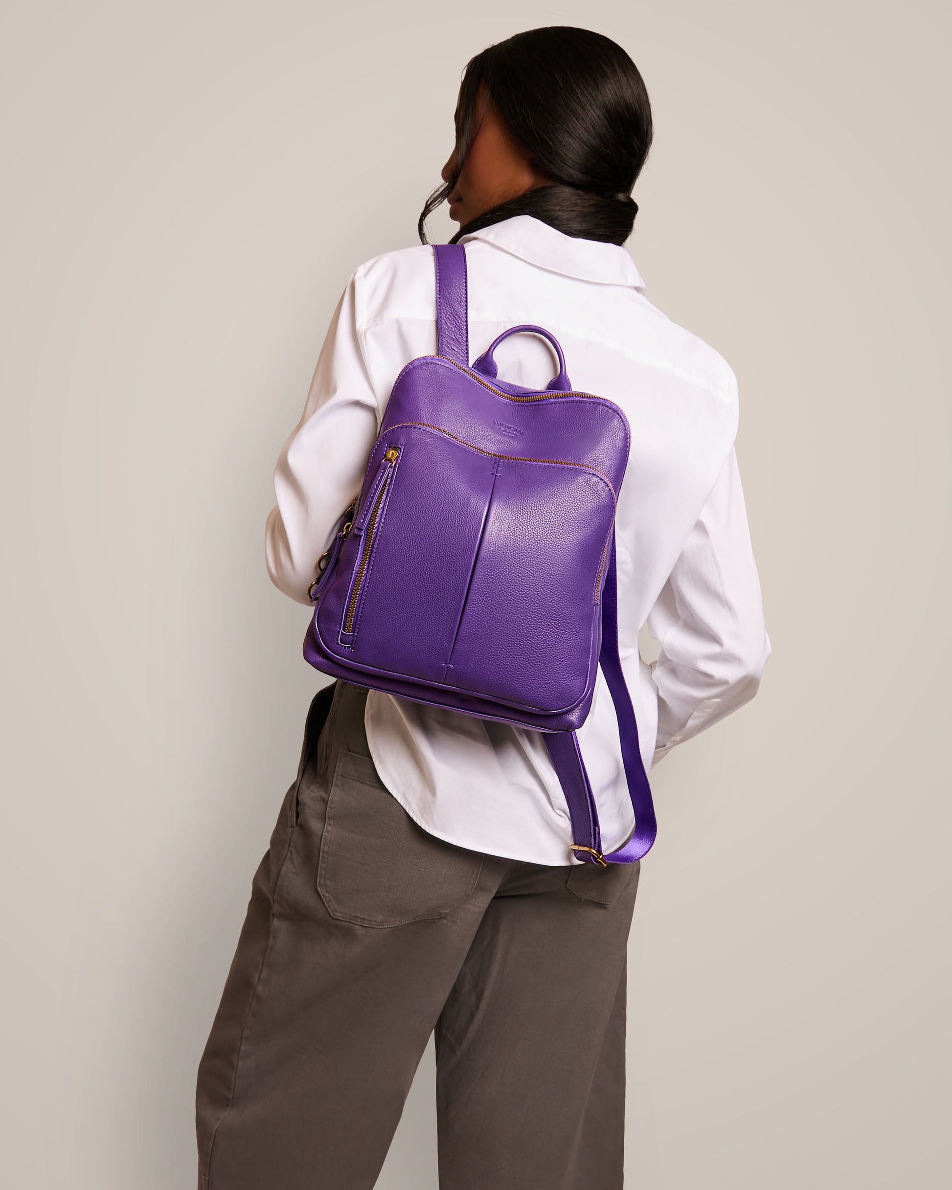 A person in a white shirt and gray pants is seen from behind, carrying the Cleveland Backpack by American Leather Co., which is a purple leather backpack purse with adjustable back straps. The plain, light-colored background highlights the rich texture of its genuine leather.