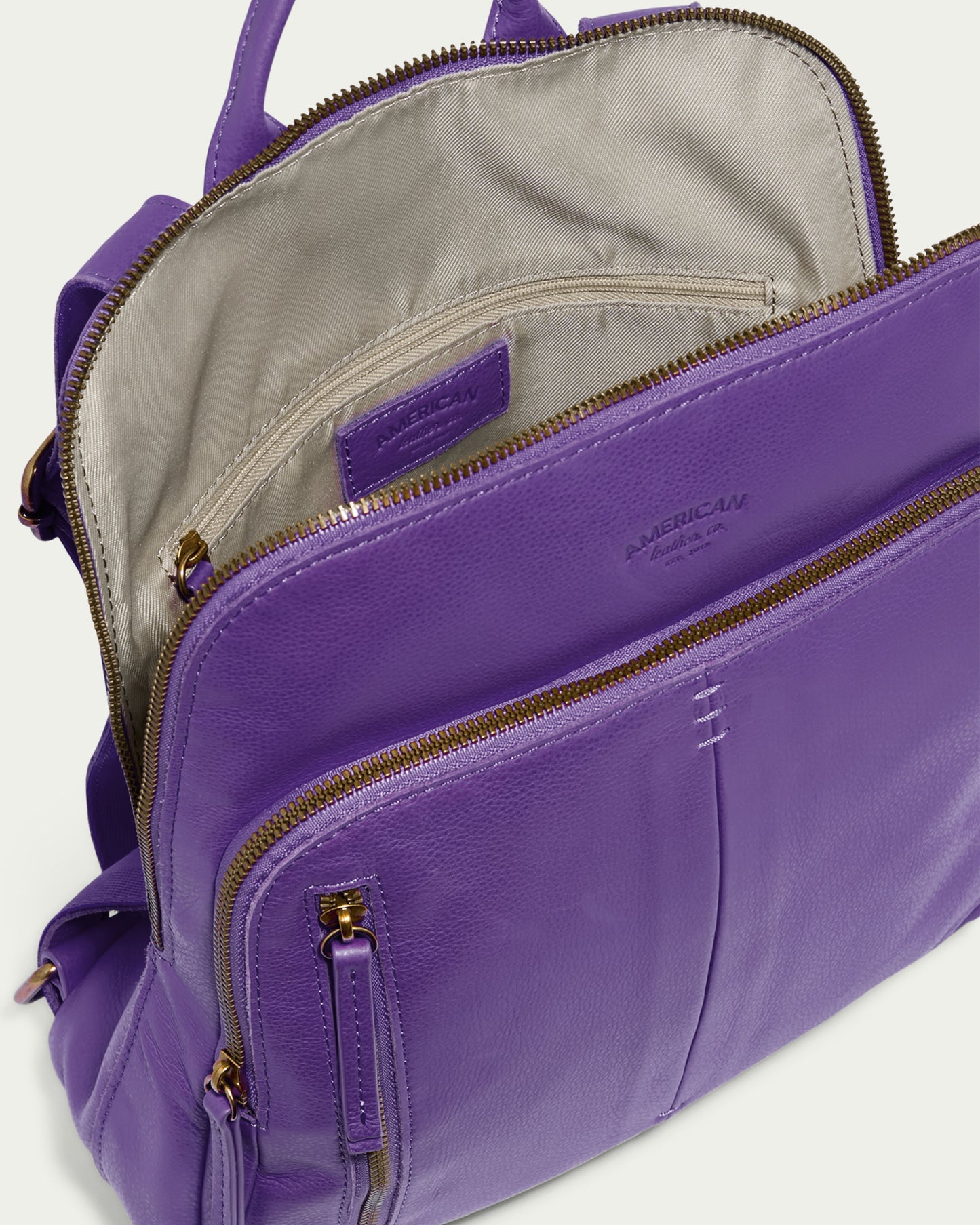 The Cleveland Backpack by American Leather Co. is a genuine leather purse in purple, featuring multiple zippered compartments. Its interior is lined with light gray fabric and equipped with organizational pockets. Gold-toned hardware enhances the zippers, and adjustable back straps offer a perfect fit.