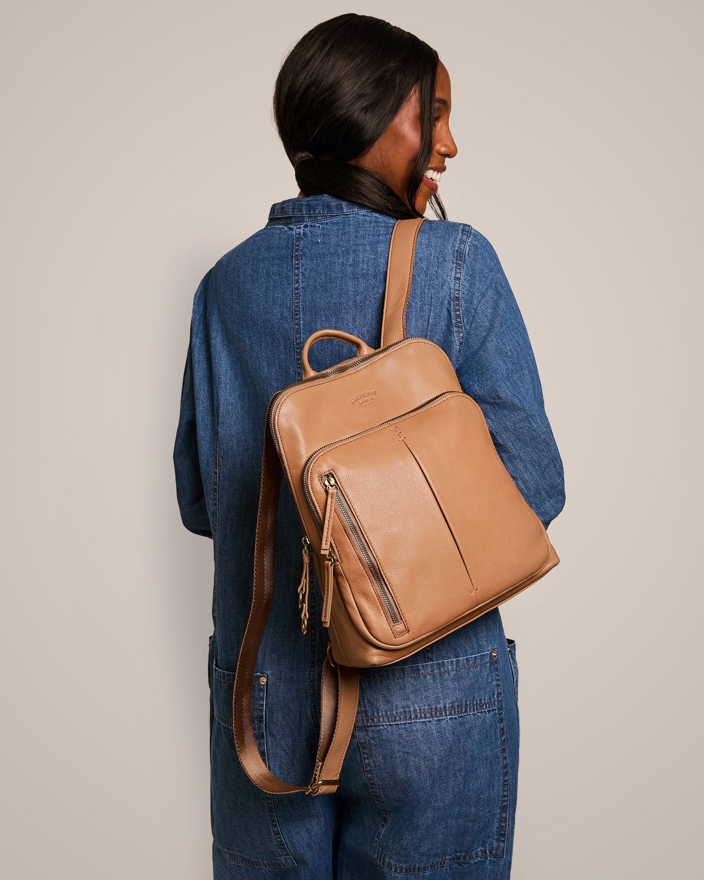 A person with long hair is wearing a denim jacket and carrying the Cleveland Backpack from American Leather Co., made of genuine leather in a tan shade. They are smiling and looking over their shoulder, set against a plain and neutral background.