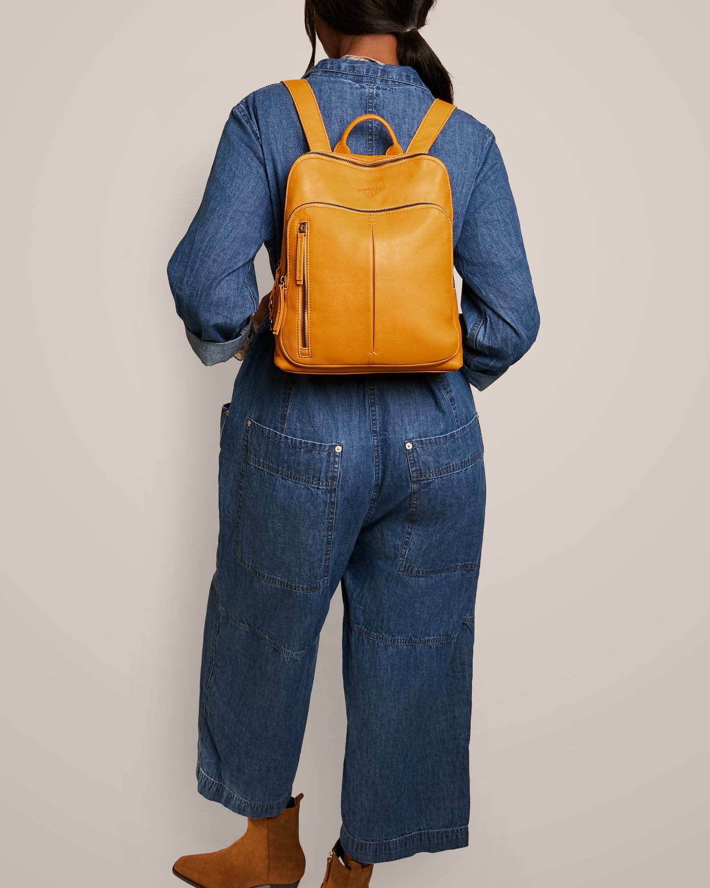 A person wearing a denim jumpsuit and brown ankle boots is facing away, sporting a stylish Cleveland Backpack by American Leather Co., crafted from genuine leather in a vibrant yellow hue.