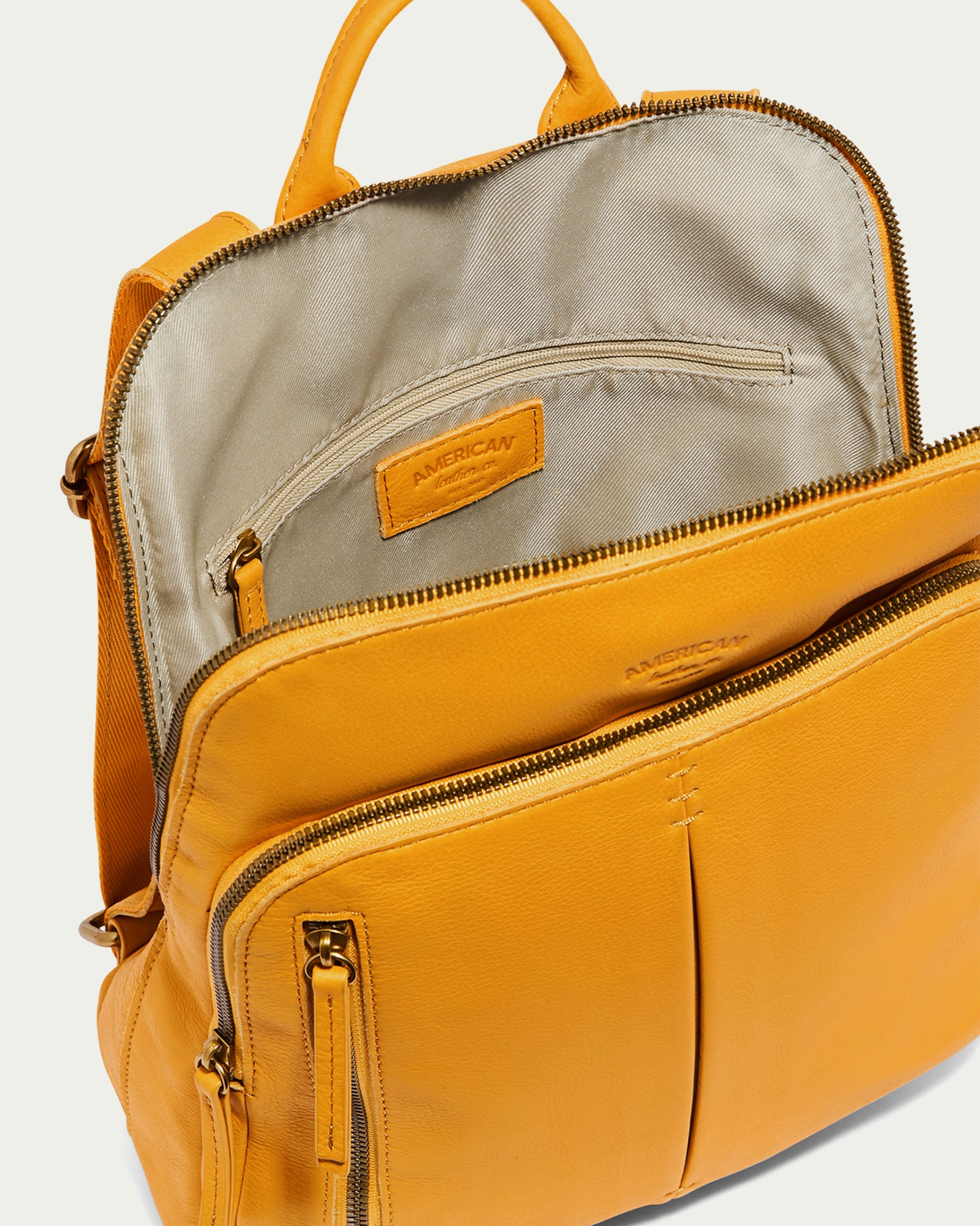 The Cleveland Backpack from American Leather Co. is displayed open, featuring an inside zipper pocket. This chic mustard yellow backpack combines functionality and elegance with its smooth genuine leather construction and two front zippered pockets.