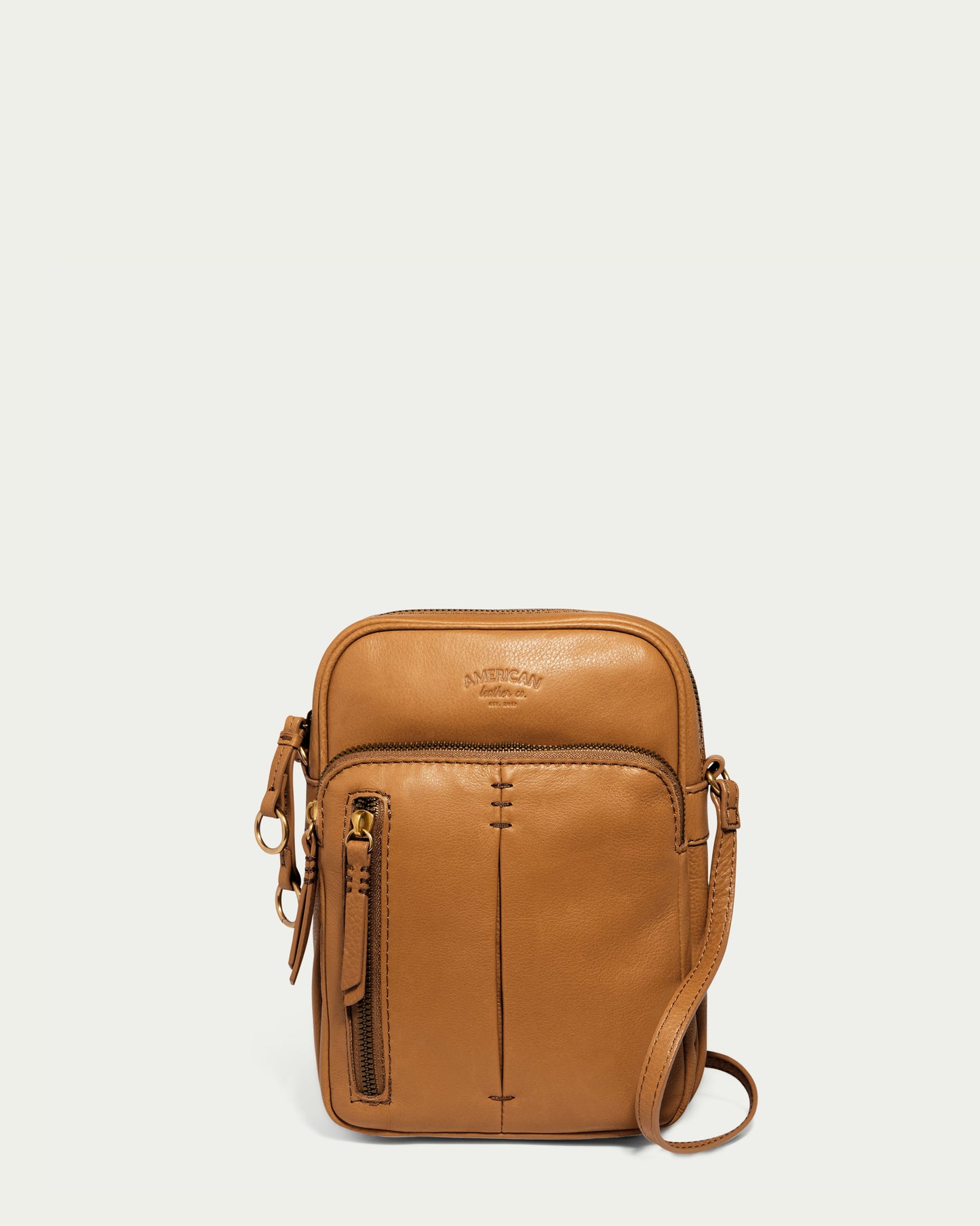 The Cleveland Large NS Crossbody by American Leather Co. is a small, tan leather bag crafted from genuine American leather with an embossed logo. It features a front zipper pocket and an adjustable crossbody strap, radiating simple elegance against a plain off-white background.