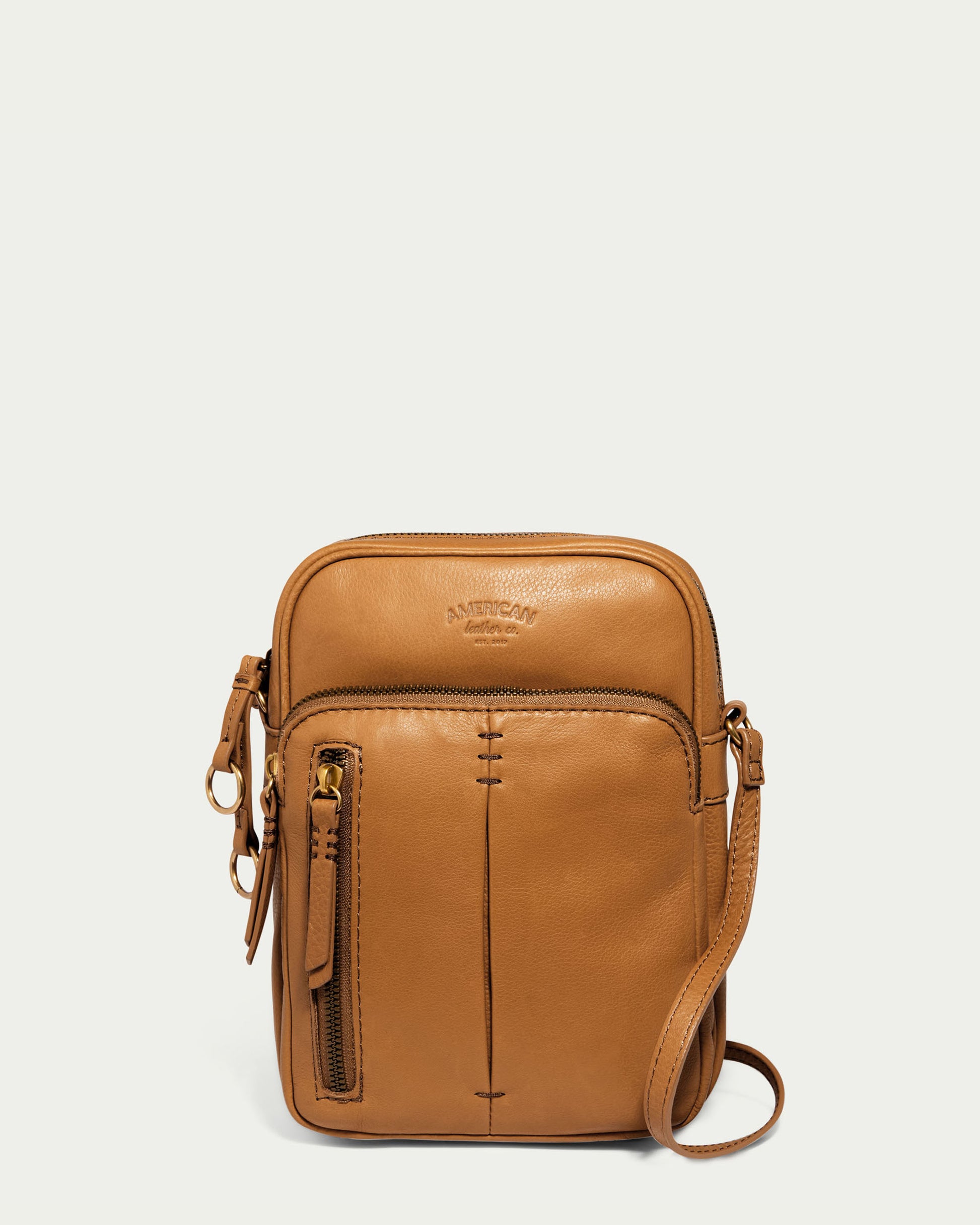 The Cleveland Large NS Crossbody by American Leather Co. is made from genuine leather and includes a front zipper pocket, small embossed logo, adjustable strap, stitched details, and gold-tone hardware. This tan bag stands out against a white background.