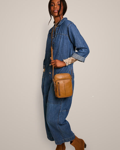 A person dressed in a denim jumpsuit and brown ankle boots is posing with a Cleveland Large NS Crossbody from American Leather Co., which boasts an adjustable crossbody strap. The ensemble is casual yet stylish, perfectly accentuated by the bag crafted from genuine American leather. The backdrop is a neutral beige.