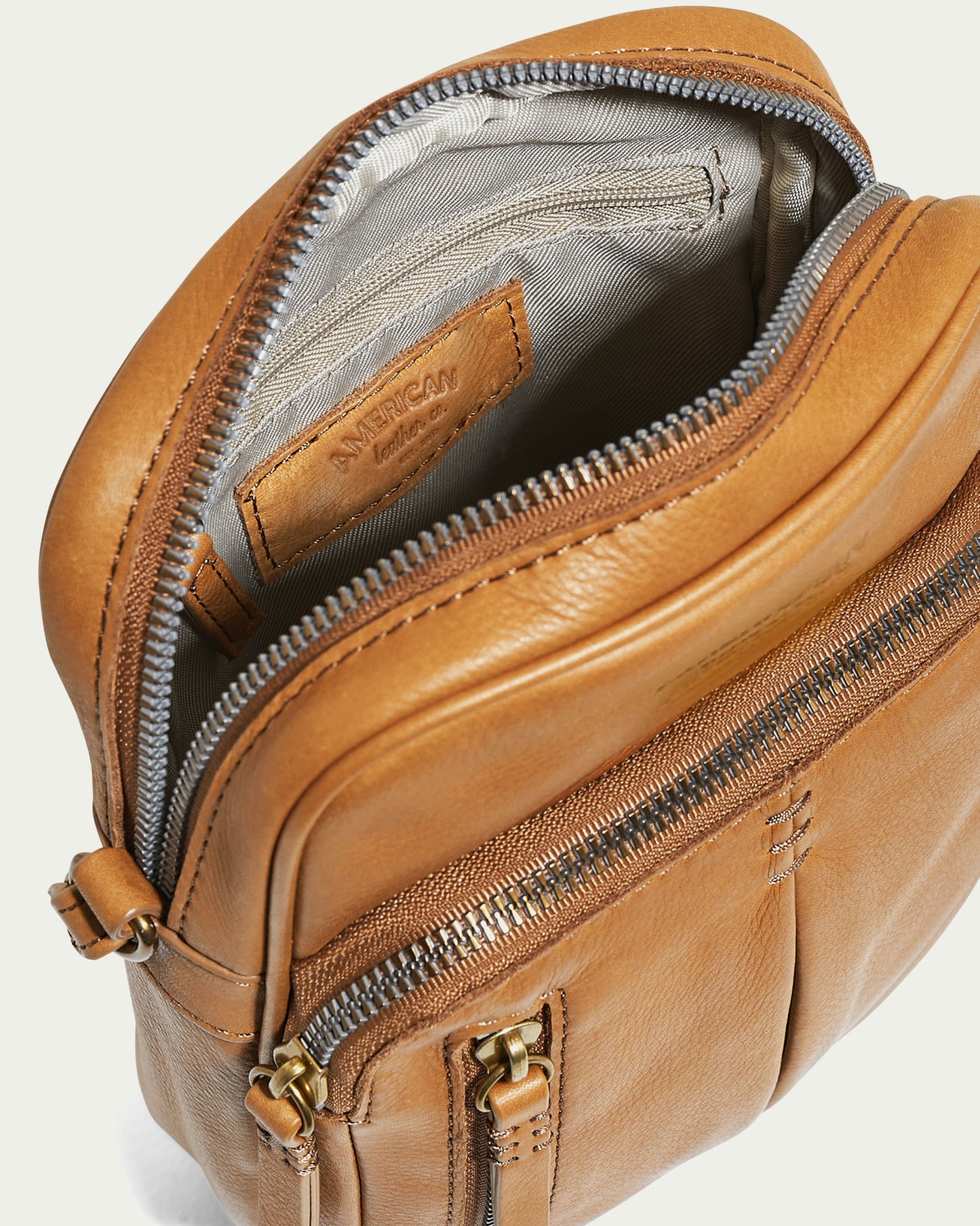 The Cleveland Large NS Crossbody by American Leather Co. is a brown leather shoulder bag made from Genuine American Leather. It includes an adjustable crossbody strap and a partially open top zipper that reveals a grey interior. The bag also features a front zipper pocket and elegant gold hardware for added style.