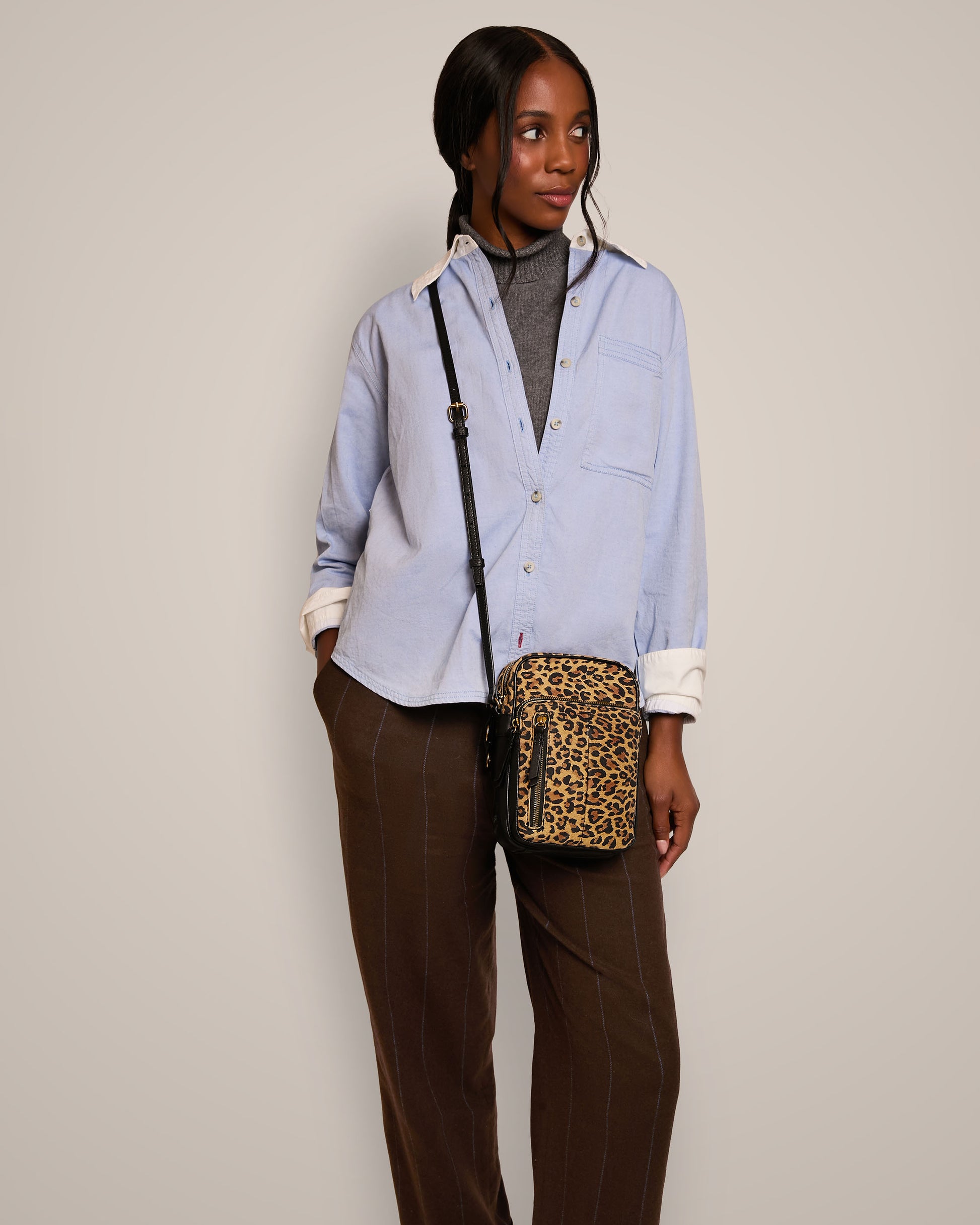 A person stands against a plain background, wearing a light blue button-up shirt and brown pants, accessorized with the Cleveland Large NS Crossbody leopard print bag from American Leather Co. Their hands are in their pockets as they look to the side.