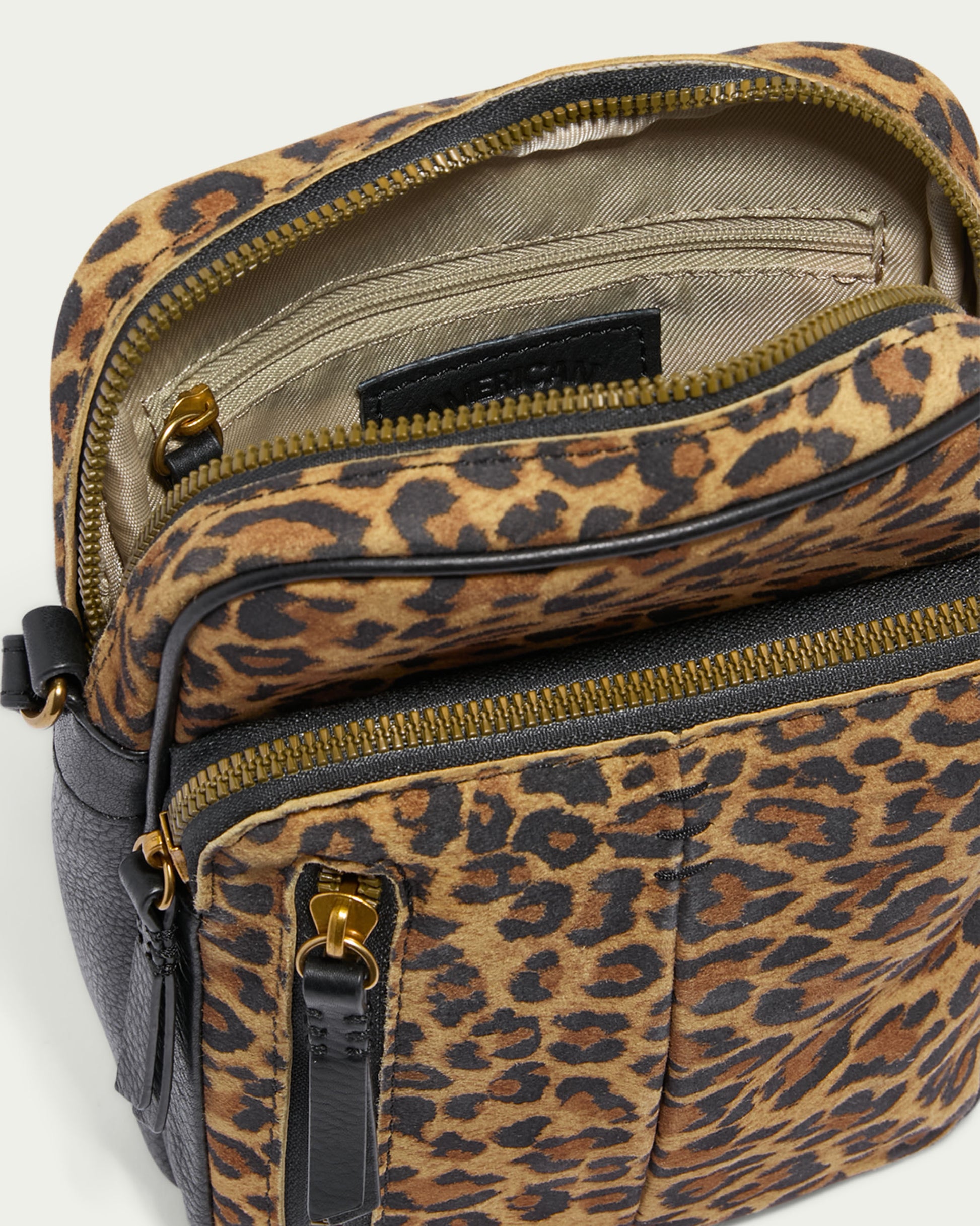 A close-up of the American Leather Co. Cleveland Large NS Crossbody reveals multiple compartments featuring gold zippers. Made from genuine leather, it boasts a leopard-print exterior with black accents. The main section and a front pocket are visible, appealing to those who value both style and functionality.