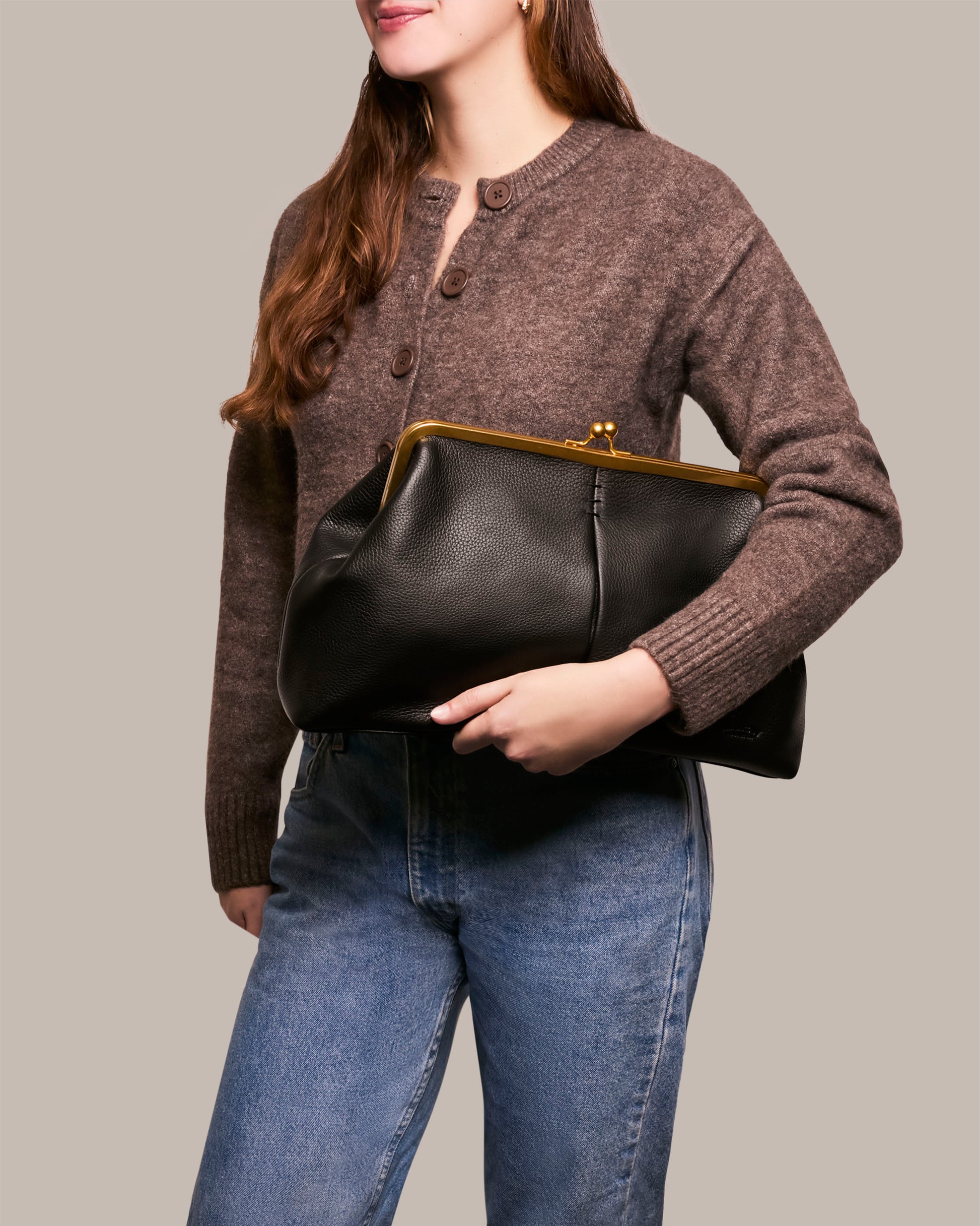 A woman in a brown sweater and blue jeans holds the Grace Oversized Clutch by American Leather Co. against a neutral background. Her long, loose hair adds elegance, while this large black leather handbag with a gold clasp features glove leather and an optional wristlet for versatile carry.