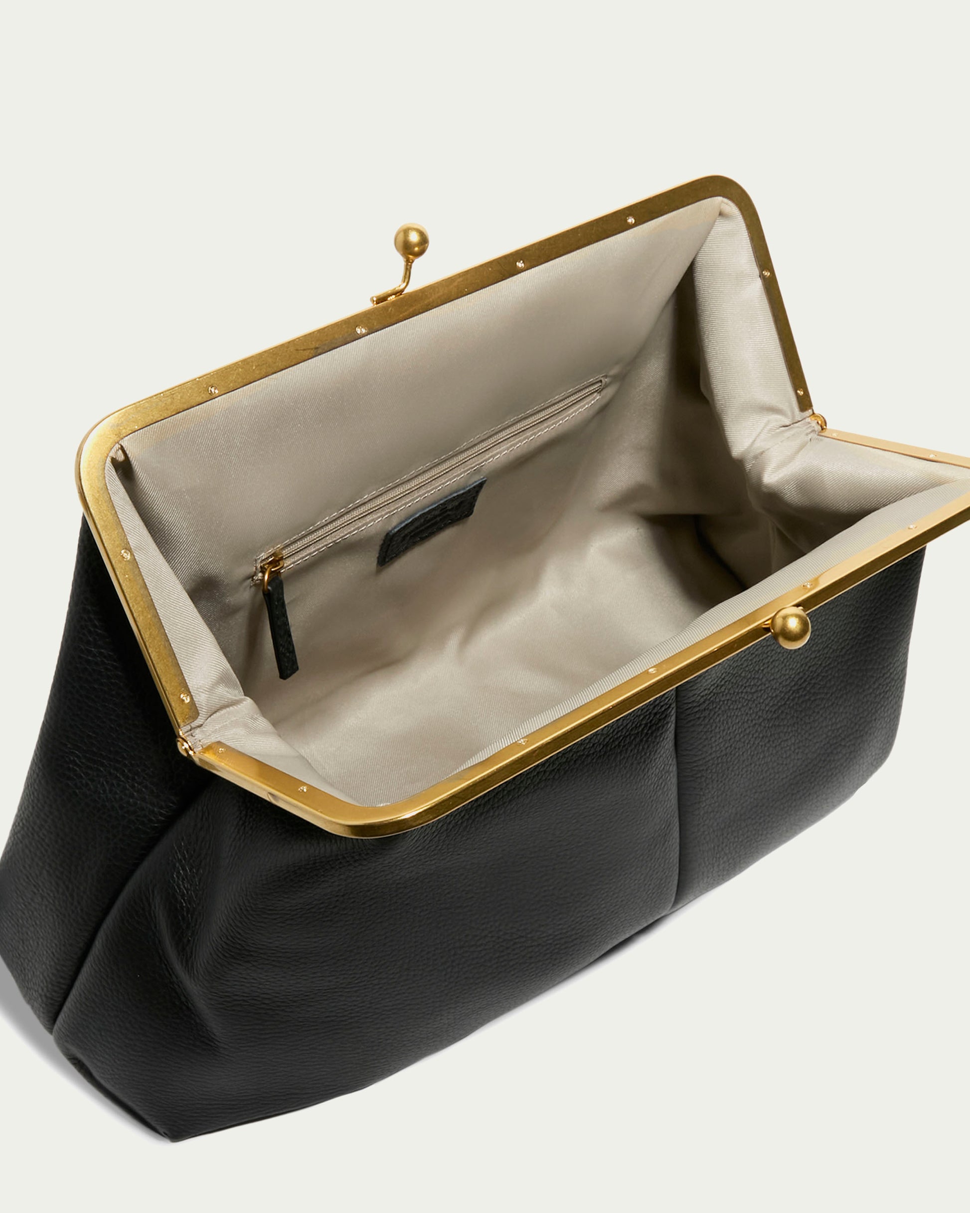 The Grace Oversized Clutch by American Leather Co. is a black textured leather handbag with a glove leather feel, beige fabric lining, gold metal frame and clasp, an interior zipper pocket, and an easy-to-use wristlet strap against a light backdrop.