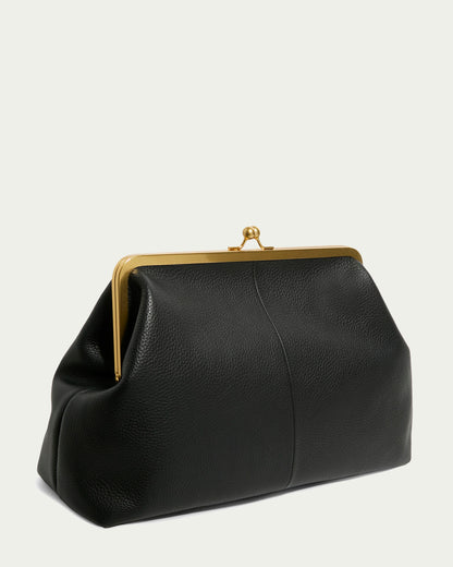 The Grace Oversized Clutch by American Leather Co. is a black leather handbag with a gold metal frame and clasp closure on a light background. It features textured glove leather, a structured triangular shape, and a convenient wristlet strap for easy carrying.