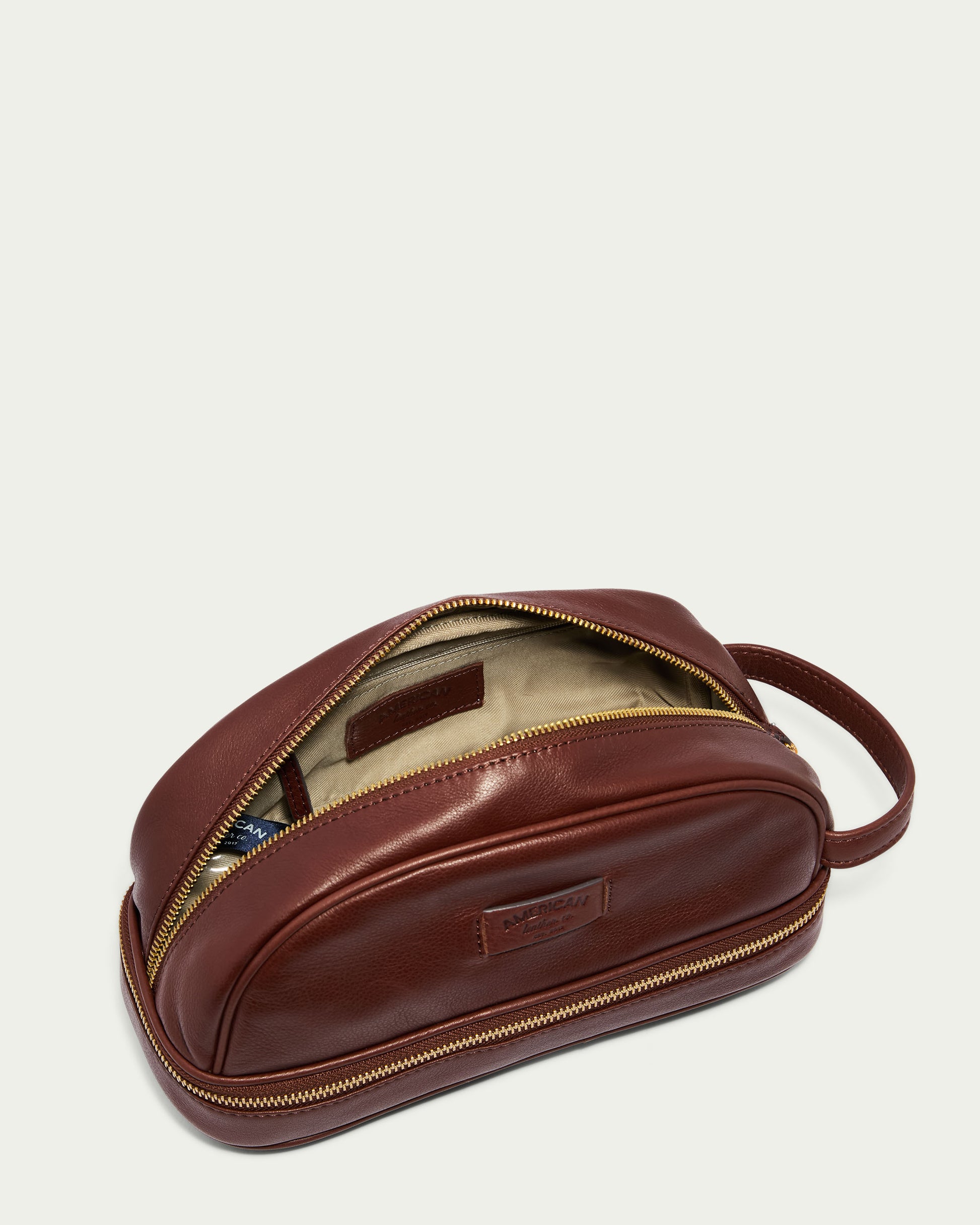 The Shorewood Dopp Kit by American Leather Co. is a brown men's glove leather dopp kit with a partially open zipper showing a beige and patterned fabric interior. It features a side handle and a rectangular brand patch on the front.