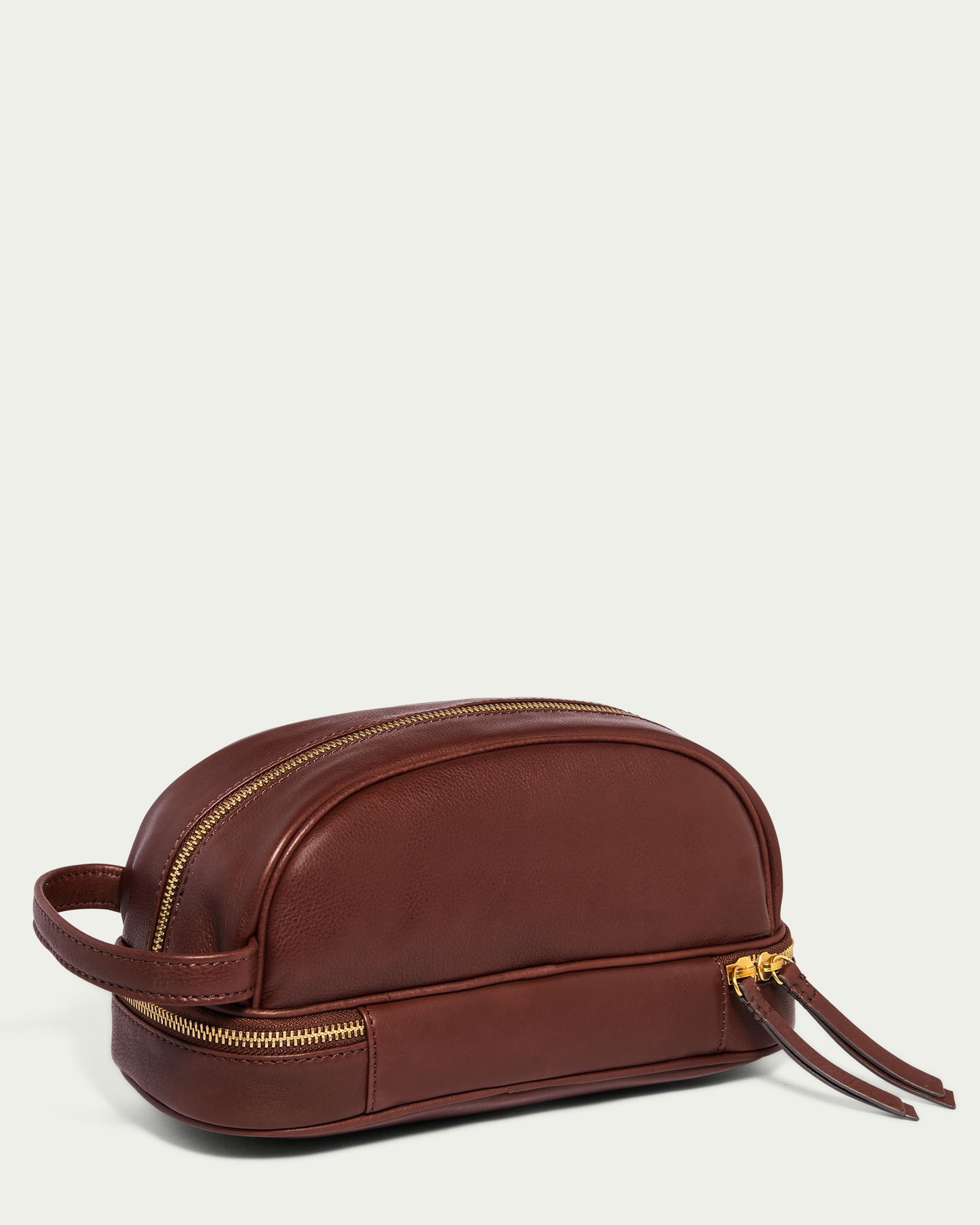 The Shorewood Dopp Kit by American Leather Co. is a men's travel bag crafted from glove leather, showcasing a brown exterior with a zipper and side handle. Its two elegant leather pull tabs and smooth, polished finish create a striking contrast against the light cream background.