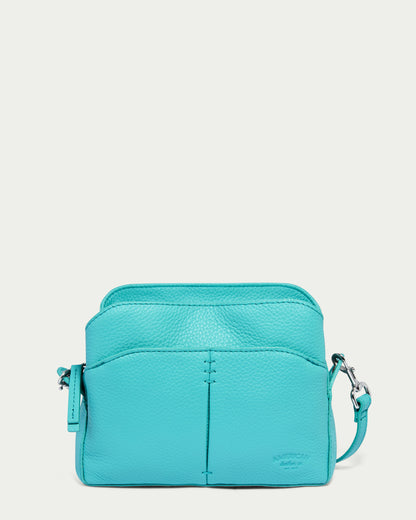 The Tulip Triple Entry Crossbody from American Leather Co. is a structured bag crafted from genuine leather in a Blue Lagoon Pebble finish. It features a front pocket with a vertical seam, a top zipper, and an adjustable strap with silver hardware, making it perfect for casual outings.