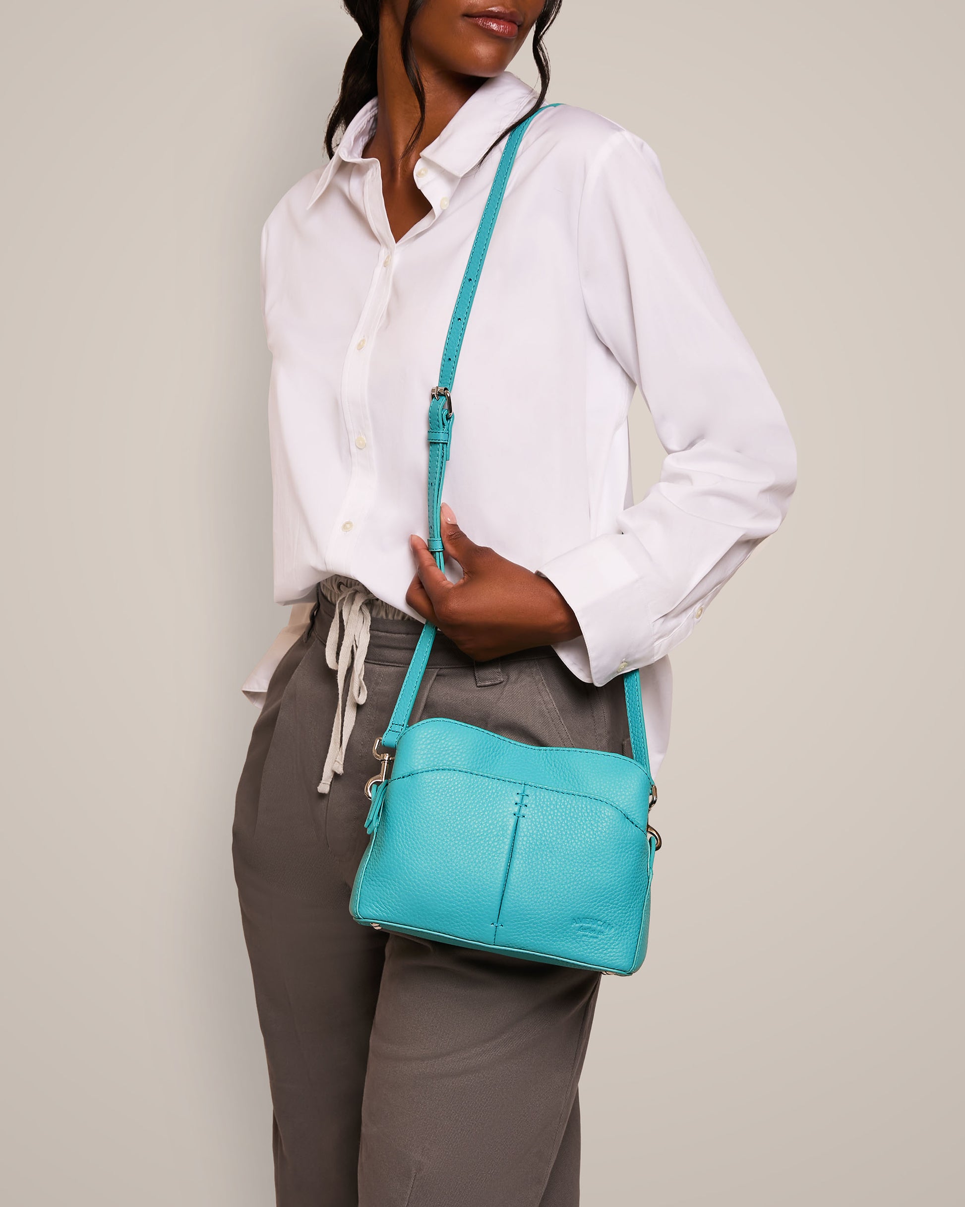 A person dressed in a white button-up shirt and gray pants is holding the Tulip Triple Entry Crossbody by American Leather Co. This turquoise accessory, made from genuine leather, features an adjustable strap and stands out beautifully against the plain background.