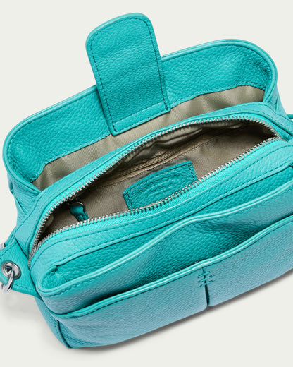 The Tulip Triple Entry Crossbody by American Leather Co. is a teal leather bag featuring a structured design and an adjustable strap for convenience. The top flap opens to reveal a beige fabric interior, complete with a zippered main compartment and an additional front pocket. Its genuine leather texture is accentuated by a silver loop for the strap attachment.