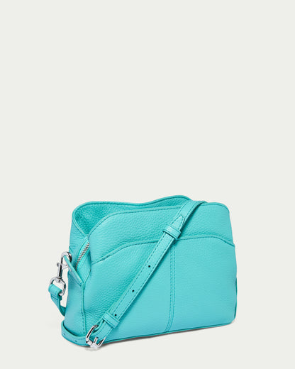 The Tulip Triple Entry Crossbody by American Leather Co. is a turquoise structured bag made from genuine leather, showcasing a silver adjustable strap and zipper. Its textured surface enhances the bag's simple, sleek design, depicted artistically at a slight angle on a white background.