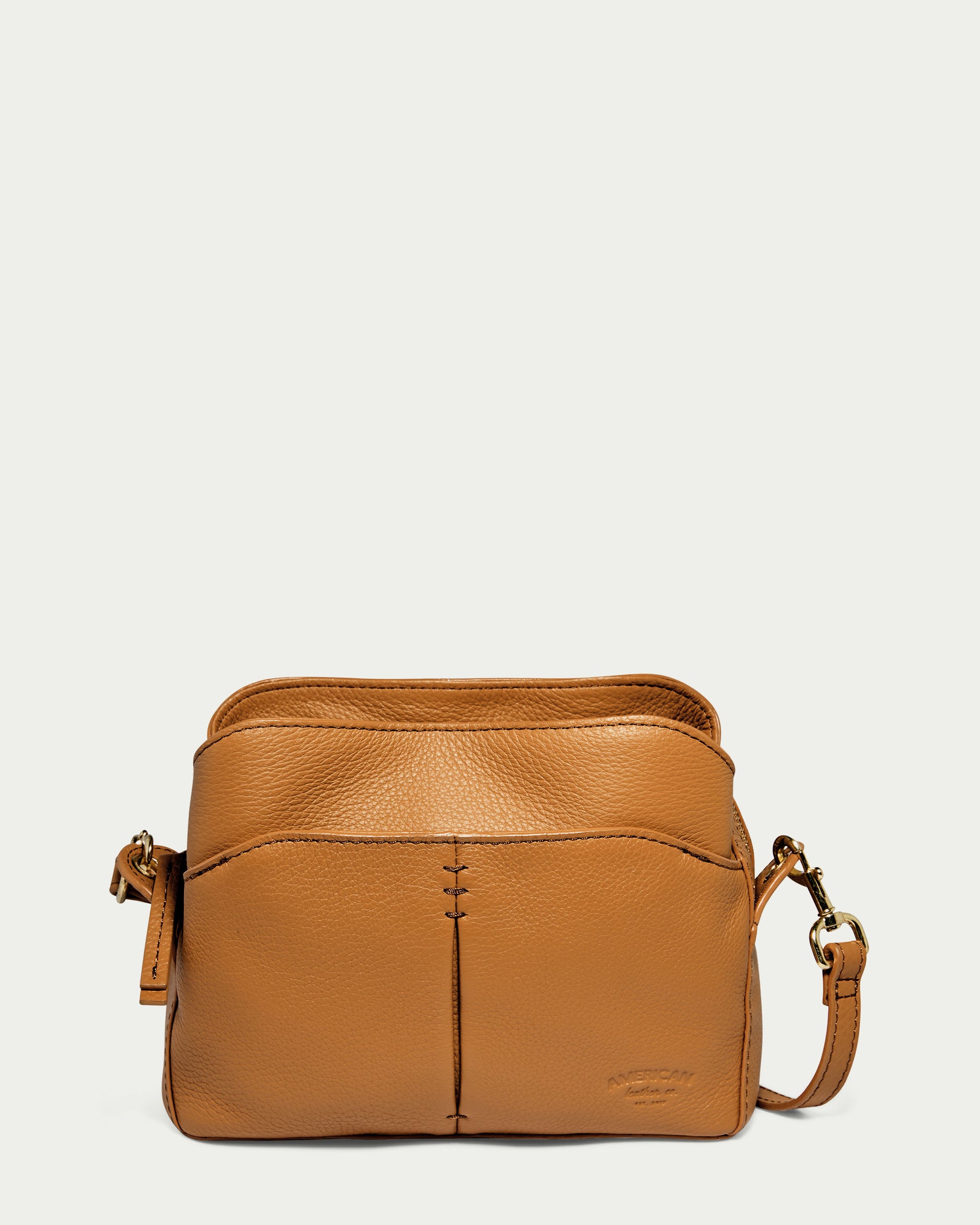 The American Leather Co. Tulip Triple Entry Crossbody in Cafe Latte Pebble is a genuine leather crossbody bag with a sleek design. It features a front pocket, zipper closure, and an adjustable strap, all while showcasing visible stitching and offering a soft texture.