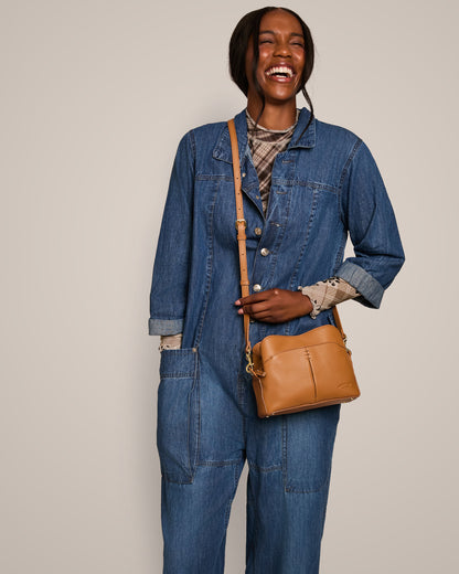 A person is standing, smiling broadly while wearing a blue denim jumpsuit over a plaid shirt, with the Tulip Triple Entry Crossbody by American Leather Co. slung over one shoulder using its adjustable strap. The background is plain and light-colored.