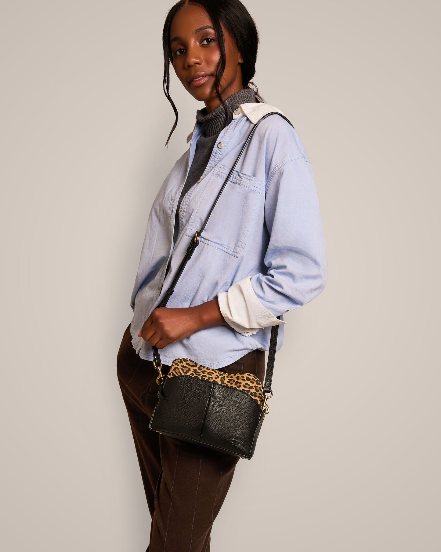 The person stands sideways, dressed in a light blue shirt layered over a gray turtleneck sweater and brown pants. They are holding the American Leather Co. Tulip Triple Entry Crossbody, featuring a genuine leather design with an adjustable gold chain strap. The background is simple and light-colored.