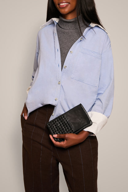 A person with long hair is wearing a light blue button-up shirt over a gray turtleneck. They hold the sleek Webster Woven Bifold Wallet by American Leather Co., which features credit card slots, and they wear brown pants with white stripes. The background is plain.