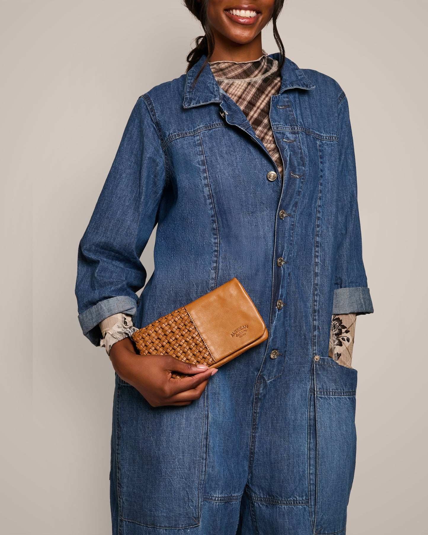 A person wearing a blue denim jumpsuit holds the American Leather Co.'s Webster Woven Bifold Wallet, crafted from supple leather. The outfit includes a patterned shirt underneath, set against a plain beige background.