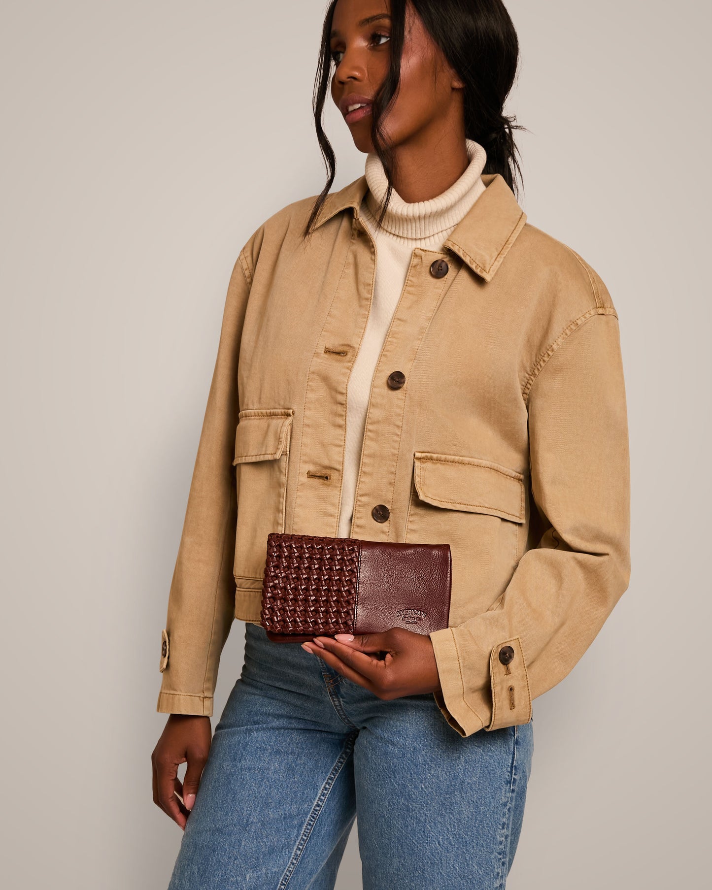 A woman in a beige jacket and blue jeans holds a maroon American Leather Co. Webster Woven Bifold Wallet. She is standing against a neutral background, with her hair styled down and wearing a light turtleneck sweater under the jacket.