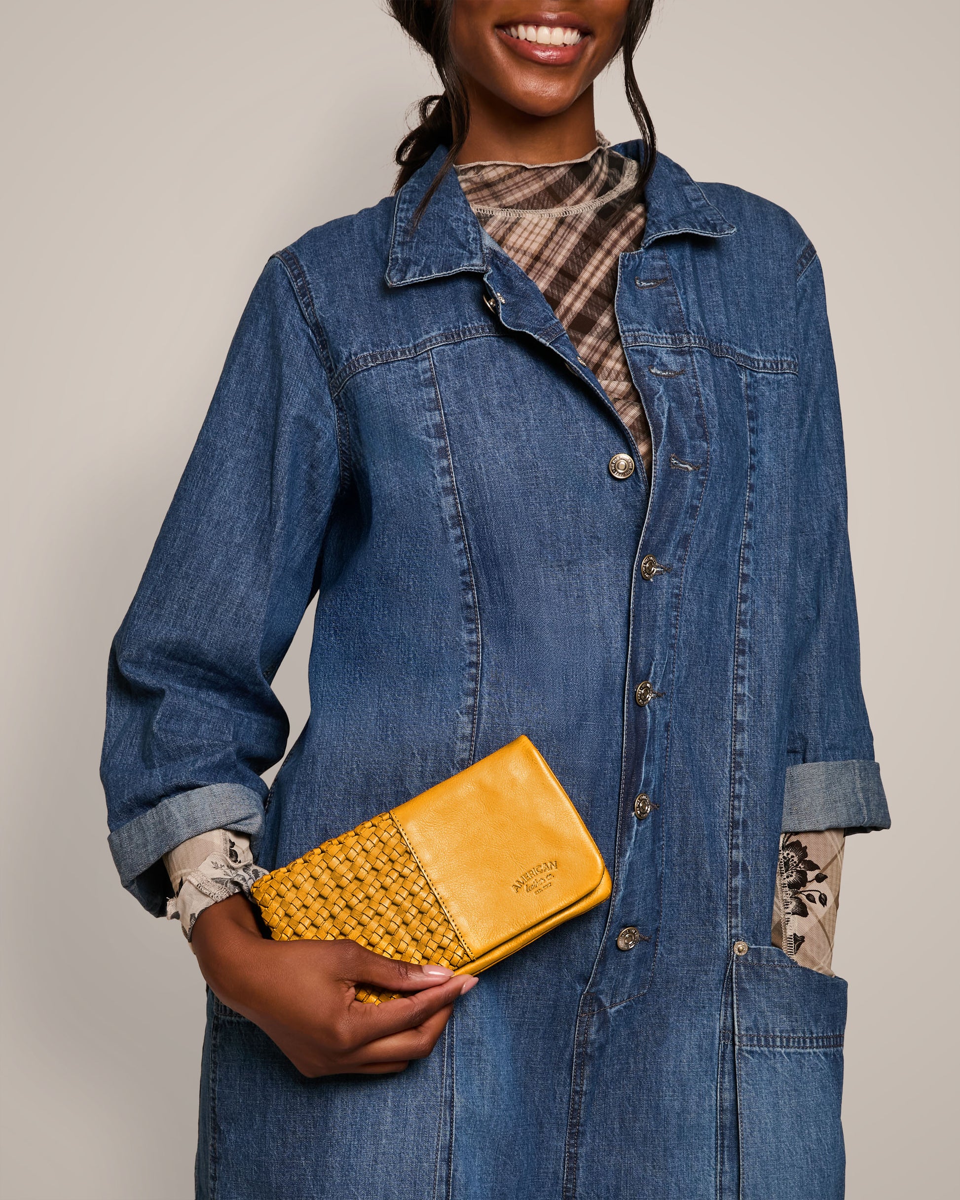 A person clad in a denim coat and plaid shirt is holding the Webster Woven Bifold Wallet from American Leather Co., featuring intricate woven details and a shiny texture. They're smiling as they showcase the yellow wallet.