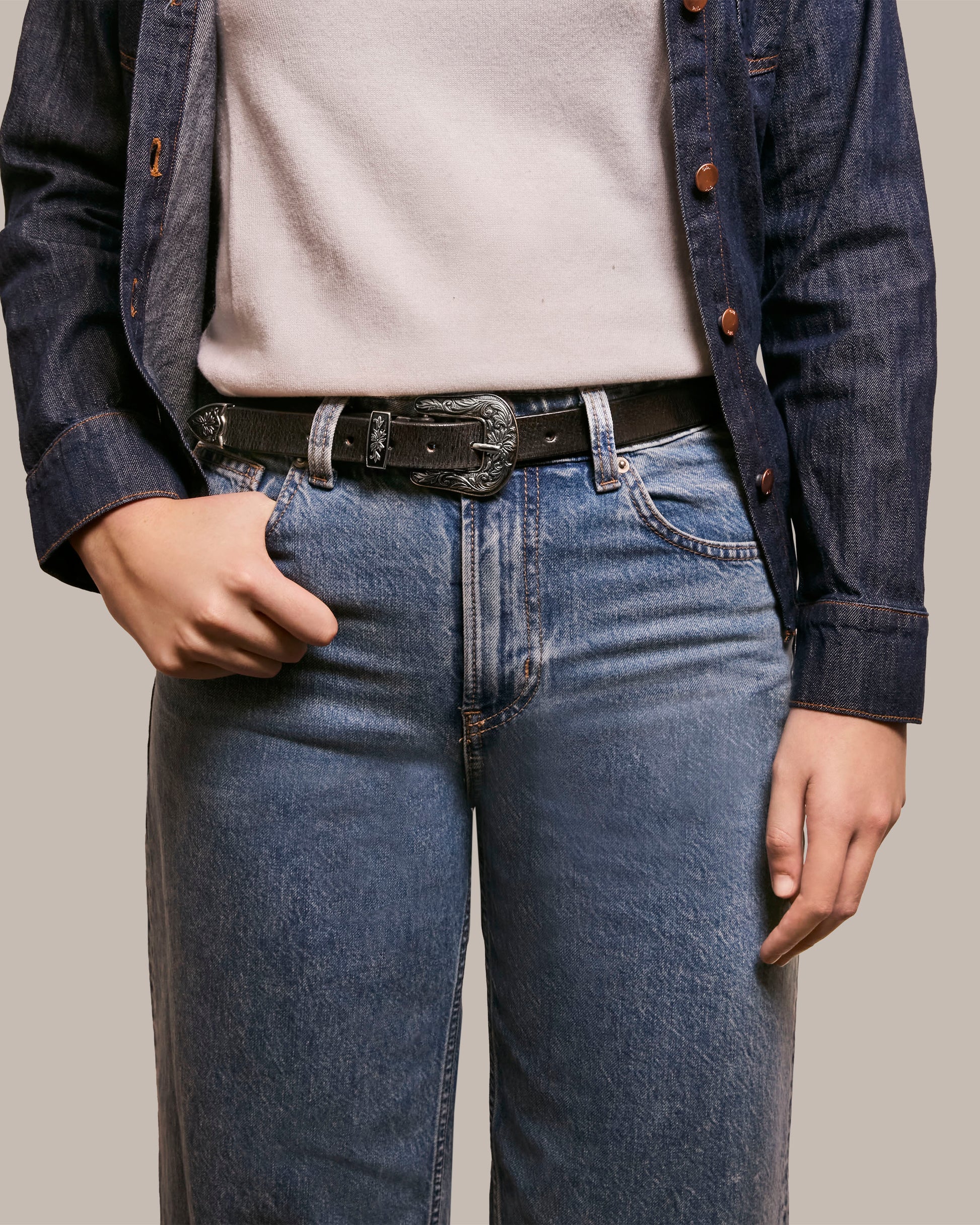 A person wearing a denim jacket, an essential wardrobe piece, over a light top and blue jeans, accessorized with the polished American Leather Co.'s Hamilton Belt, stands against a plain background.