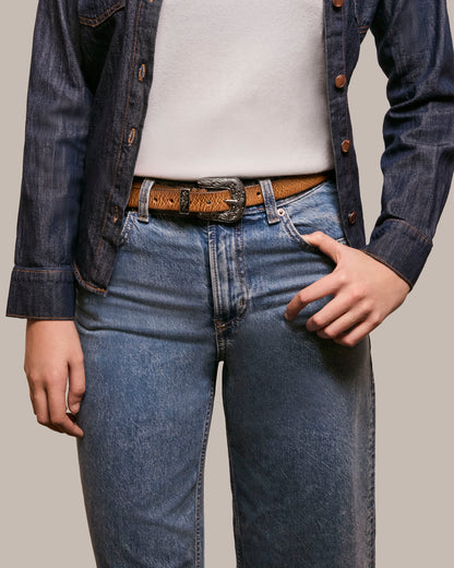 A person wearing a dark denim jacket over a white shirt, with blue jeans and the Hamilton Belt by American Leather Co. featuring polished hardware stands with one hand in their pocket against a plain background.