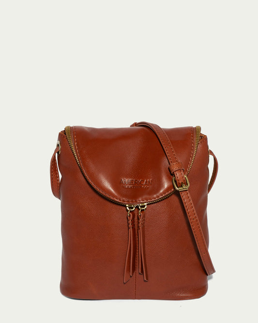 The Hampton Large Zip-Around Crossbody from American Leather Co. is a brown leather bag made from genuine glove leather, featuring a flap closure and gold zipper. It includes an adjustable crossbody strap and tassel accents, with a smooth texture and the American Leather Co. brand name embossed on the front.