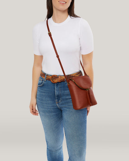 A person wearing a white t-shirt and blue jeans with a brown belt stands holding an American Leather Co. Hampton Large Zip-Around Crossbody bag crafted from Genuine Glove Leather. The background is a plain, light grey color.