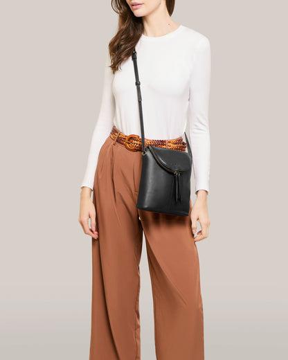 A woman is dressed in a white long-sleeve top and brown wide-leg pants, complemented by a woven belt and the Hampton Large Zip-Around Crossbody by American Leather Co., featuring an adjustable strap and tassel. Her hair flows freely as she stands against a light gray background.