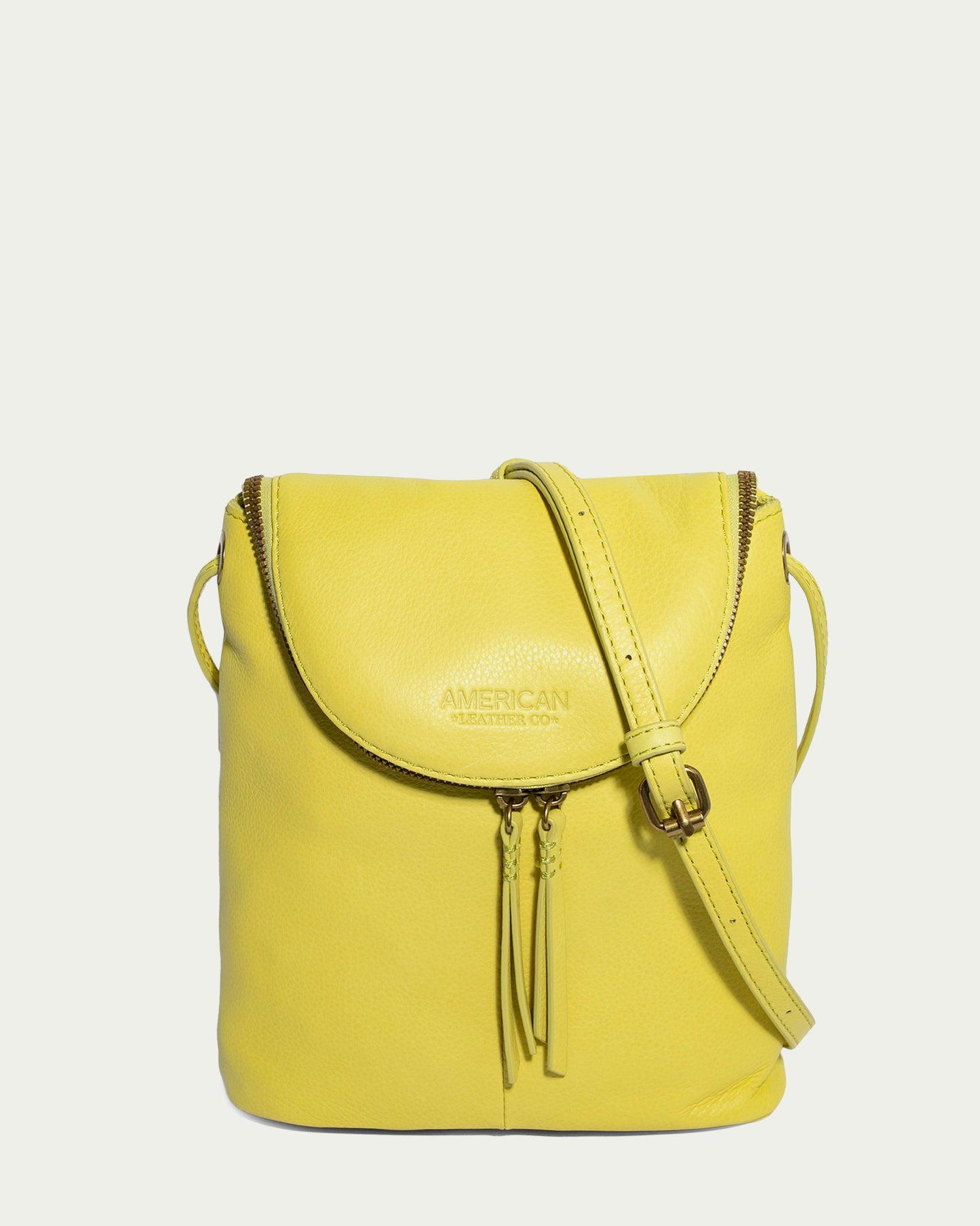 Introducing the Hampton Large Zip-Around Crossbody, a striking accessory from American Leather Co. This bag is made from genuine glove leather in bright yellow, boasting an adjustable strap and intricate tassel zipper accents. The brand's signature embossing graces the flap against a light cream backdrop.