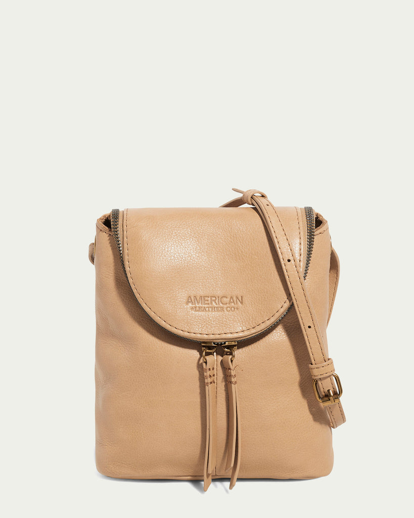 Introducing the Hampton Large Zip-Around Crossbody from American Leather Co.: This tan leather crossbody bag showcases a flap closure and is elegantly adorned with two tassels. It features an adjustable strap and is expertly crafted from genuine glove leather. The embossed text "AMERICAN LEATHER CO." graces its front, all set against a plain off-white background.