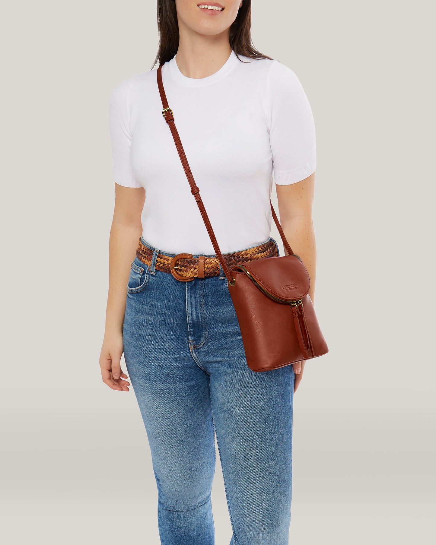 A person in a white t-shirt and blue jeans stands against a plain background, showcasing the Hampton Large Zip-Around Crossbody by American Leather Co. Crafted from genuine glove leather, this stylish bag includes a braided belt, front zipper pocket, and an adjustable crossbody strap for optimal comfort and fit.