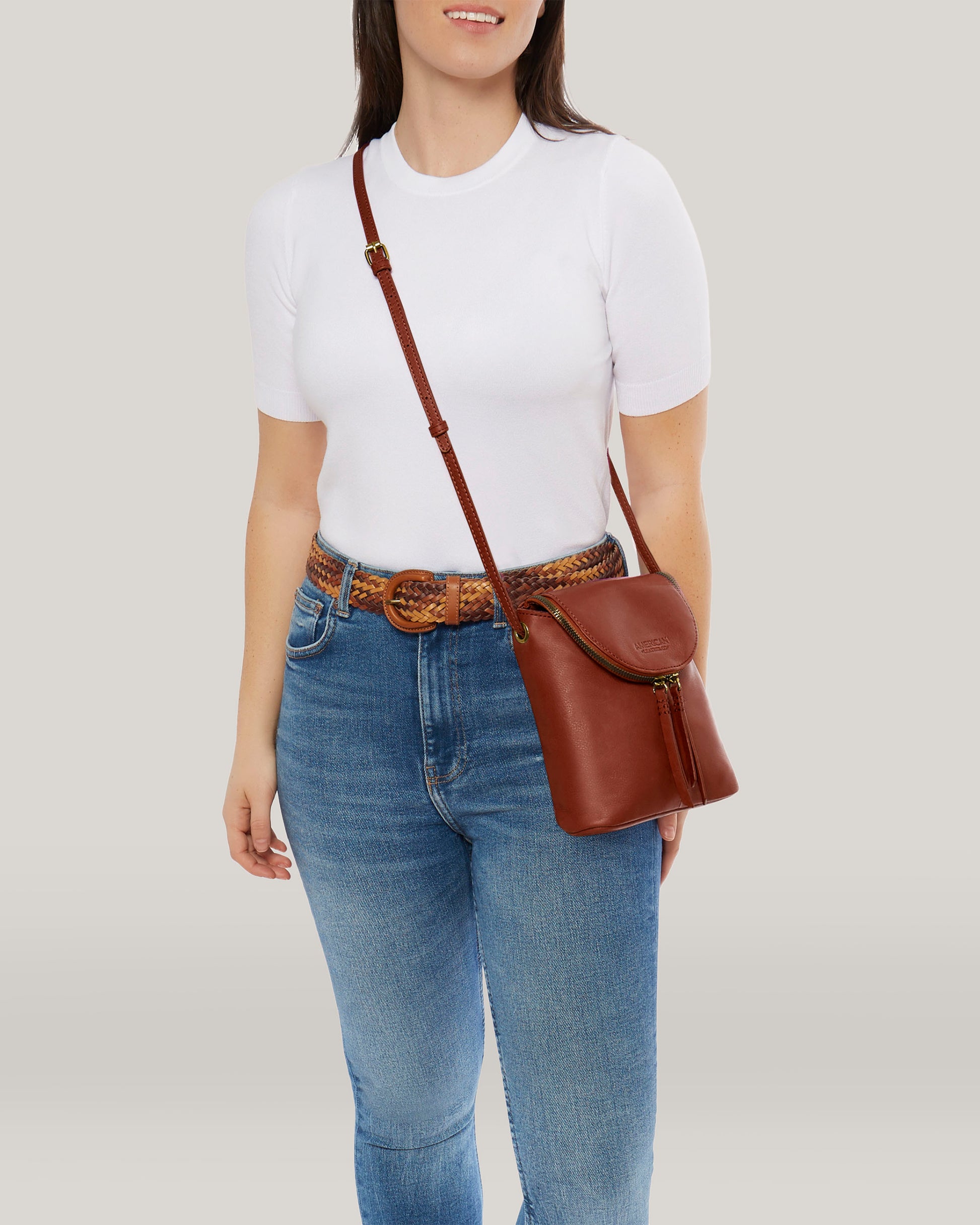 A person stands wearing a white t-shirt, blue jeans, and a woven belt. They carry an American Leather Co. Hampton Large Zip-Around Crossbody crafted from genuine glove leather, its adjustable strap perfectly complementing the ensemble. The background is a plain, light color.