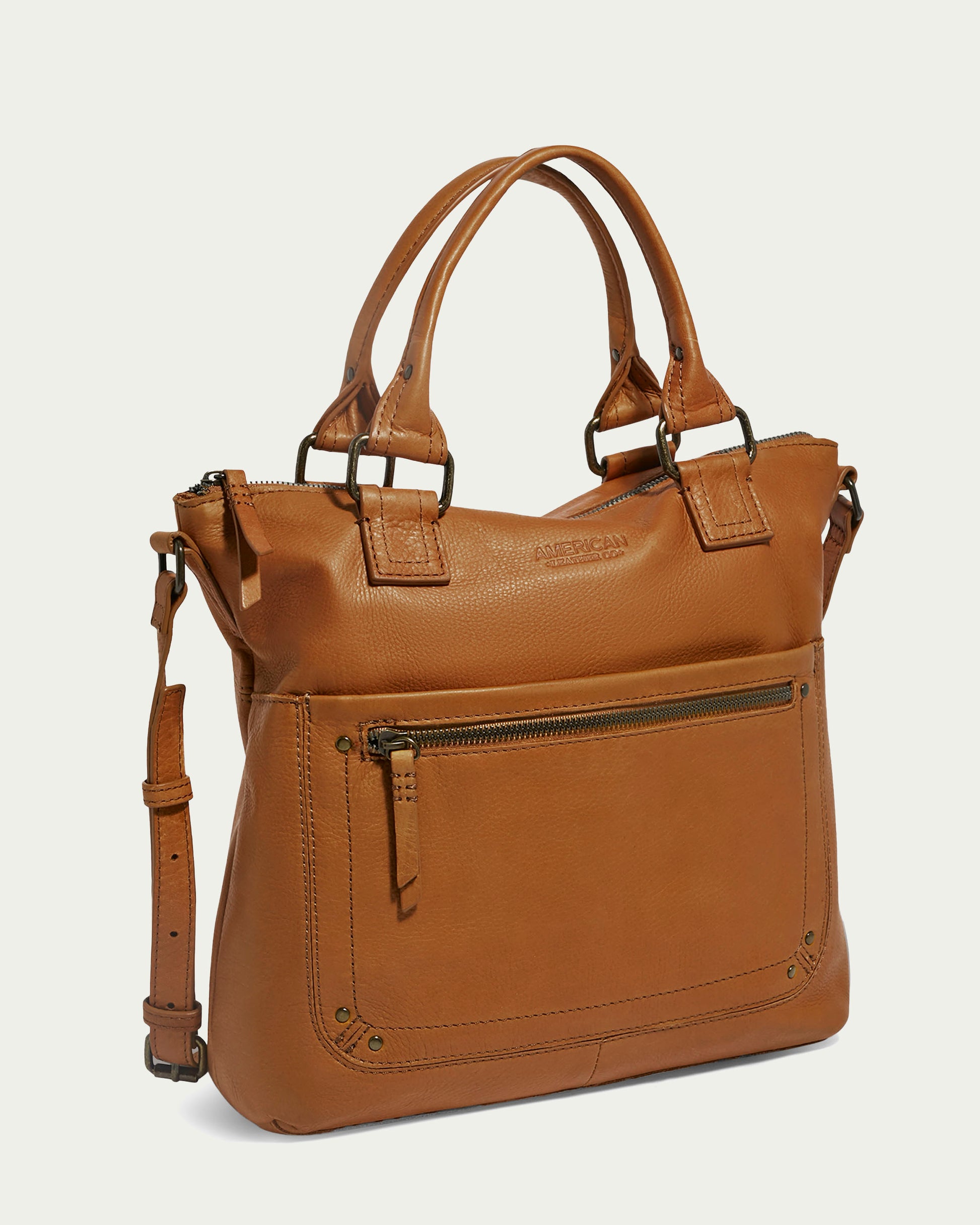 Introducing the Jamestown Tote by American Leather Co.: A sophisticated brown handbag designed from Genuine American Leather. It features dual handles, an adjustable shoulder strap, and a secure top zip closure. Adorned with metallic bronze accents, this bag includes a front zipper pocket for added convenience, elevated against a light beige backdrop.