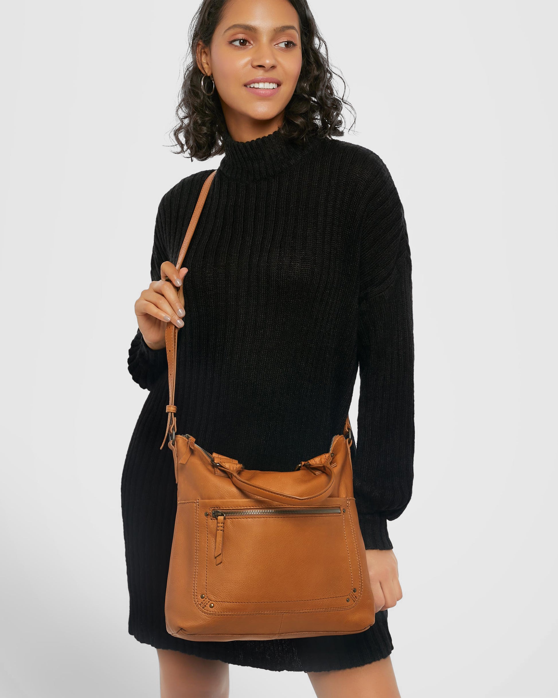 A woman with curly hair is wearing a black sweater dress and holding the Jamestown Tote from American Leather Co., a tan leather crossbody bag crafted from genuine American leather that features multiple zippered compartments and an adjustable shoulder strap. The background is plain white.