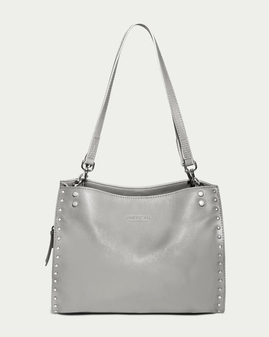 A chic Lenox Studded Triple Entry Satchel from American Leather Co. in a light gray hue with a shoulder strap and fashionable metal studs along the edges. Made from Genuine American Leather, it showcases a subtle logo in the center and includes a small zipper pocket on the side. The plain, light-colored background complements its design.