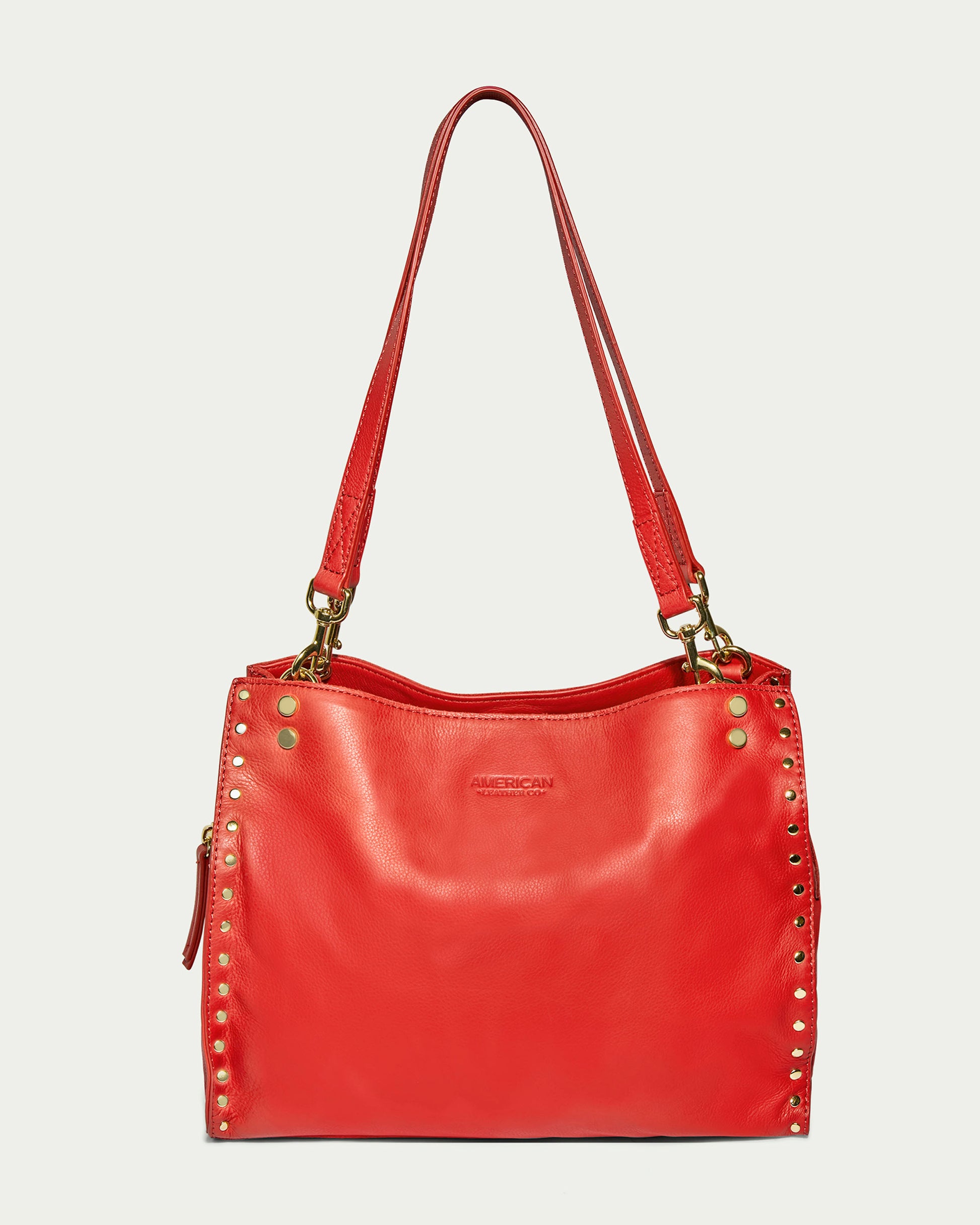 The Lenox Studded Triple Entry Satchel by American Leather Co. is a red, genuine leather handbag adorned with gold studs in a decorative pattern along the edges. It includes two shoulder straps and a zippered closure at the top. The bag boasts a sleek, polished look and features a subtle brand logo on the front.