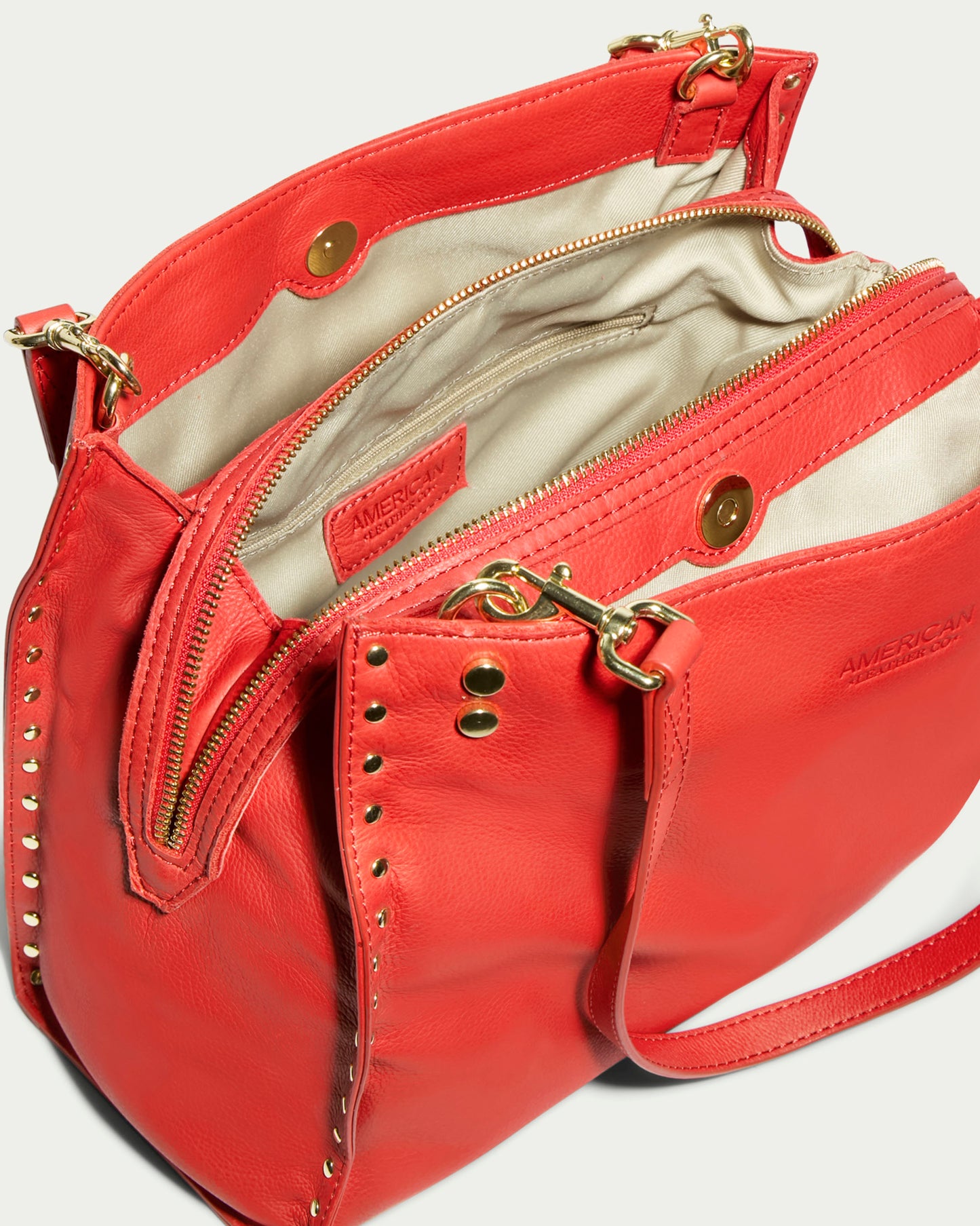 The Lenox Studded Triple Entry Satchel by American Leather Co. is a striking red handbag, crafted from genuine leather and adorned with gold hardware and studs. Its beige interior features zipper and button compartments, while a long shoulder strap enhances its functionality.