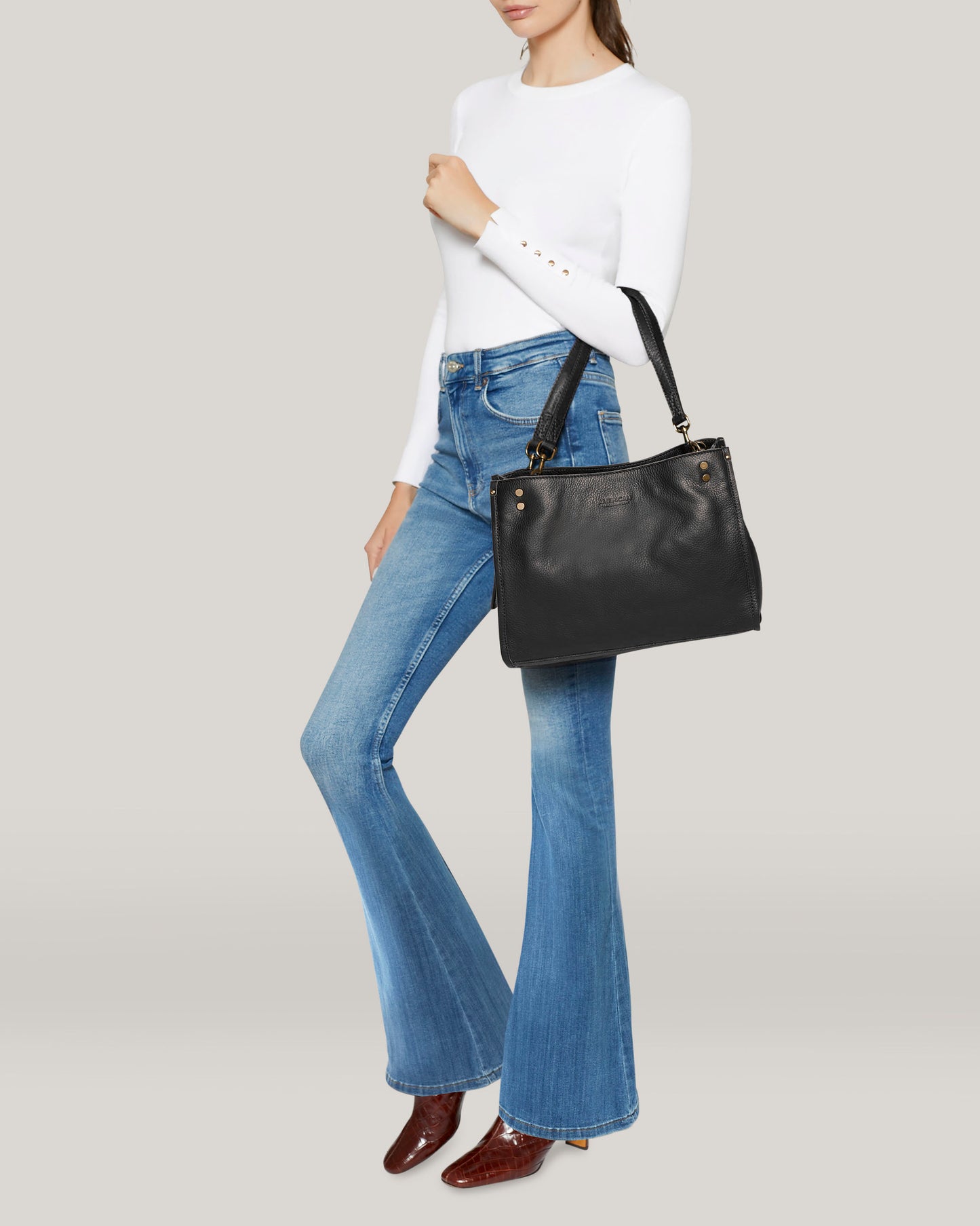 A person wearing a white long-sleeve top, blue flared jeans, and brown ankle boots holds the elegant Lenox Triple Entry Satchel by American Leather Co., crafted from genuine American leather with two straps. The background is light gray.