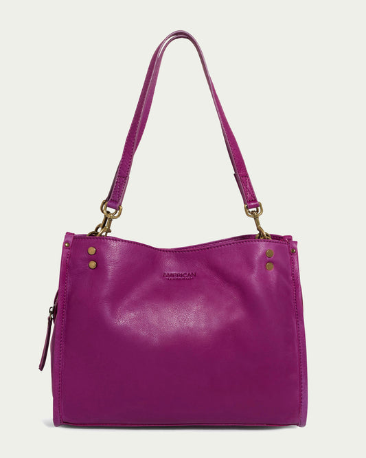 Introducing the Lenox Triple Entry Satchel from American Leather Co., a chic and vibrant accessory crafted from genuine magenta leather. Adorned with gold hardware and a convenient shoulder strap, this satchel includes a zippered side pocket and "American Leather Co." embossed on the front, making it an essential for everyday use.